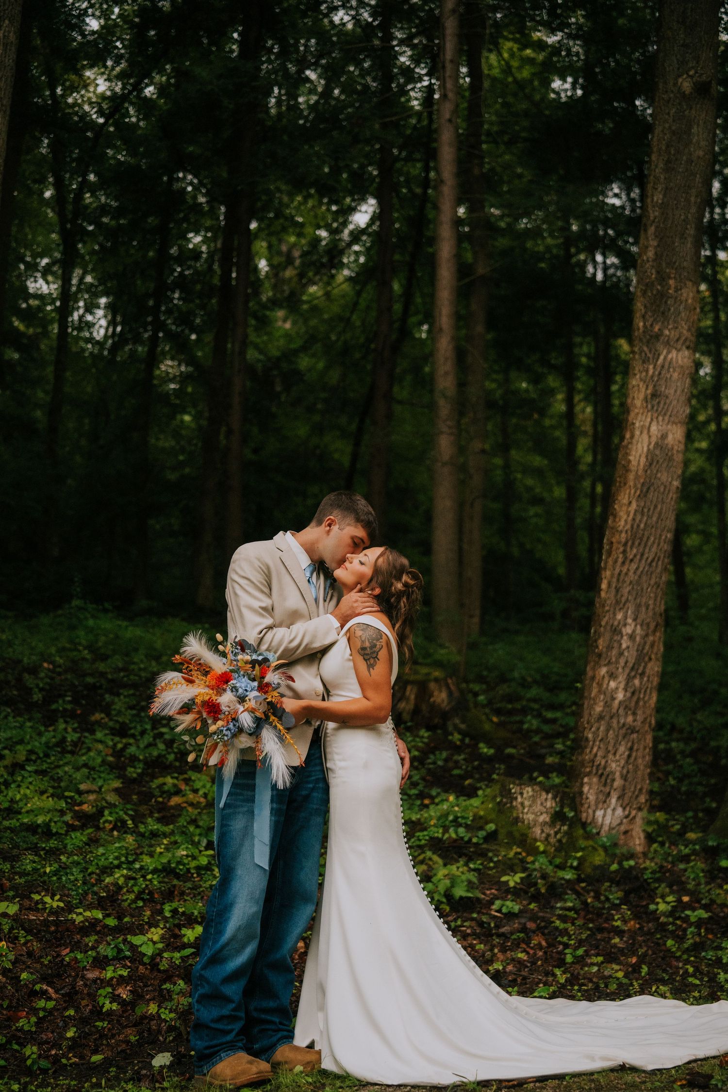 A romantic wedding photo of a couple embracing in a wooded forest setting with the bride holding a colorful bouquet.