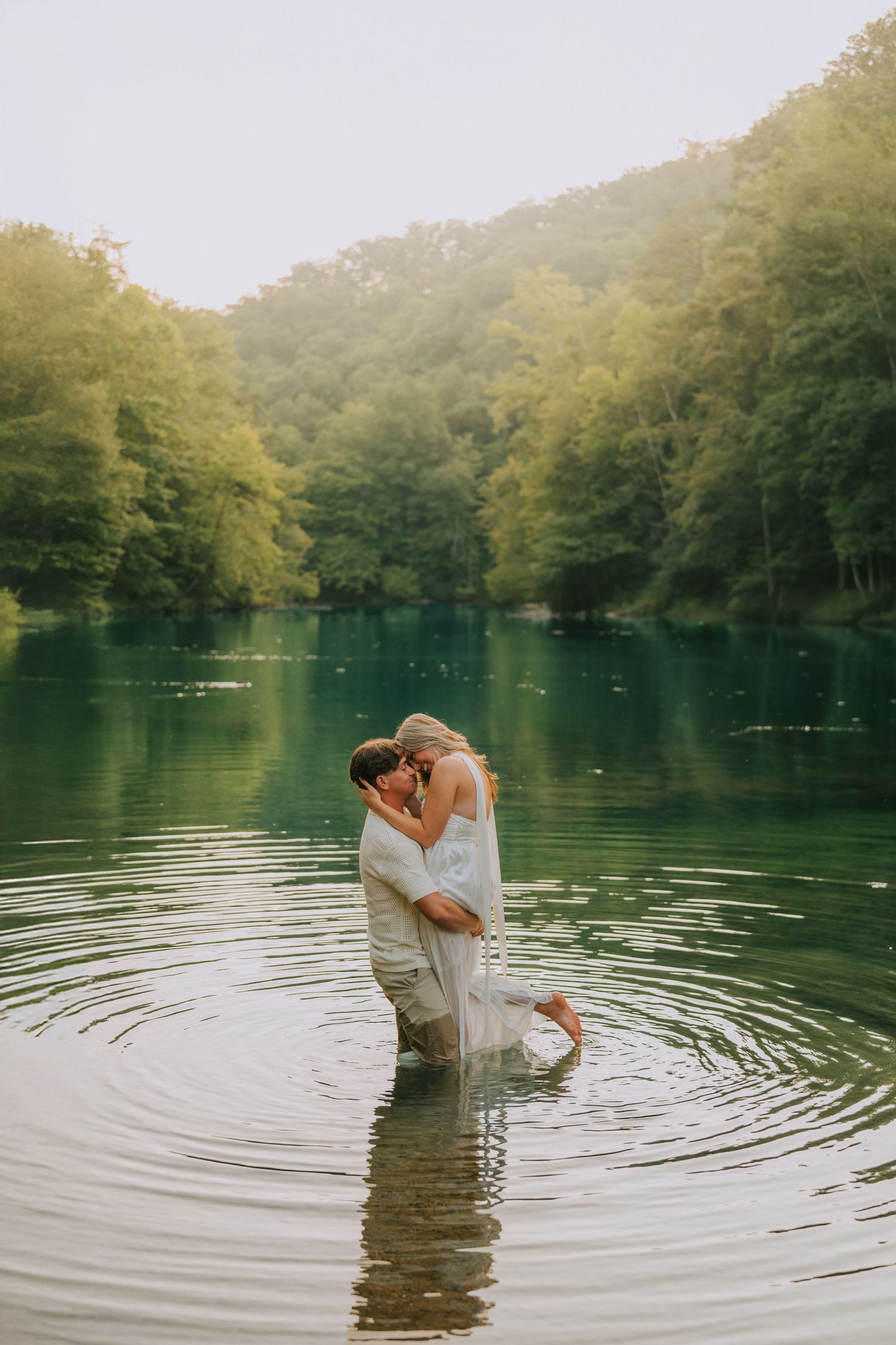 Romantic couple standing in clear lake water creating ripples during intimate photoshoot.