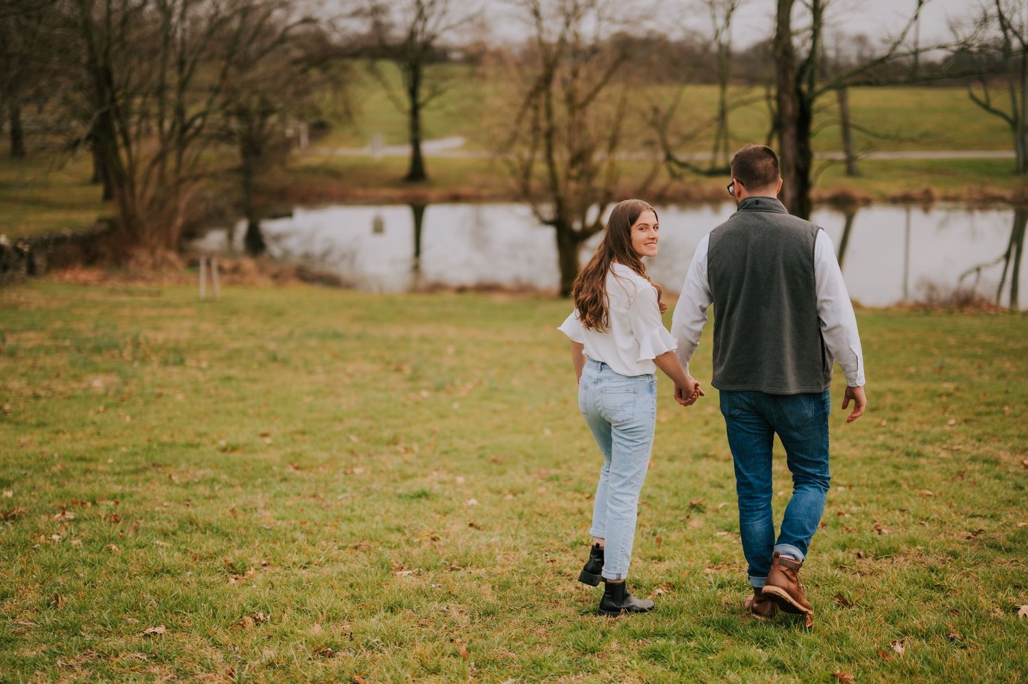 A couple walks hand in hand through a scenic outdoor setting with trees and grass.