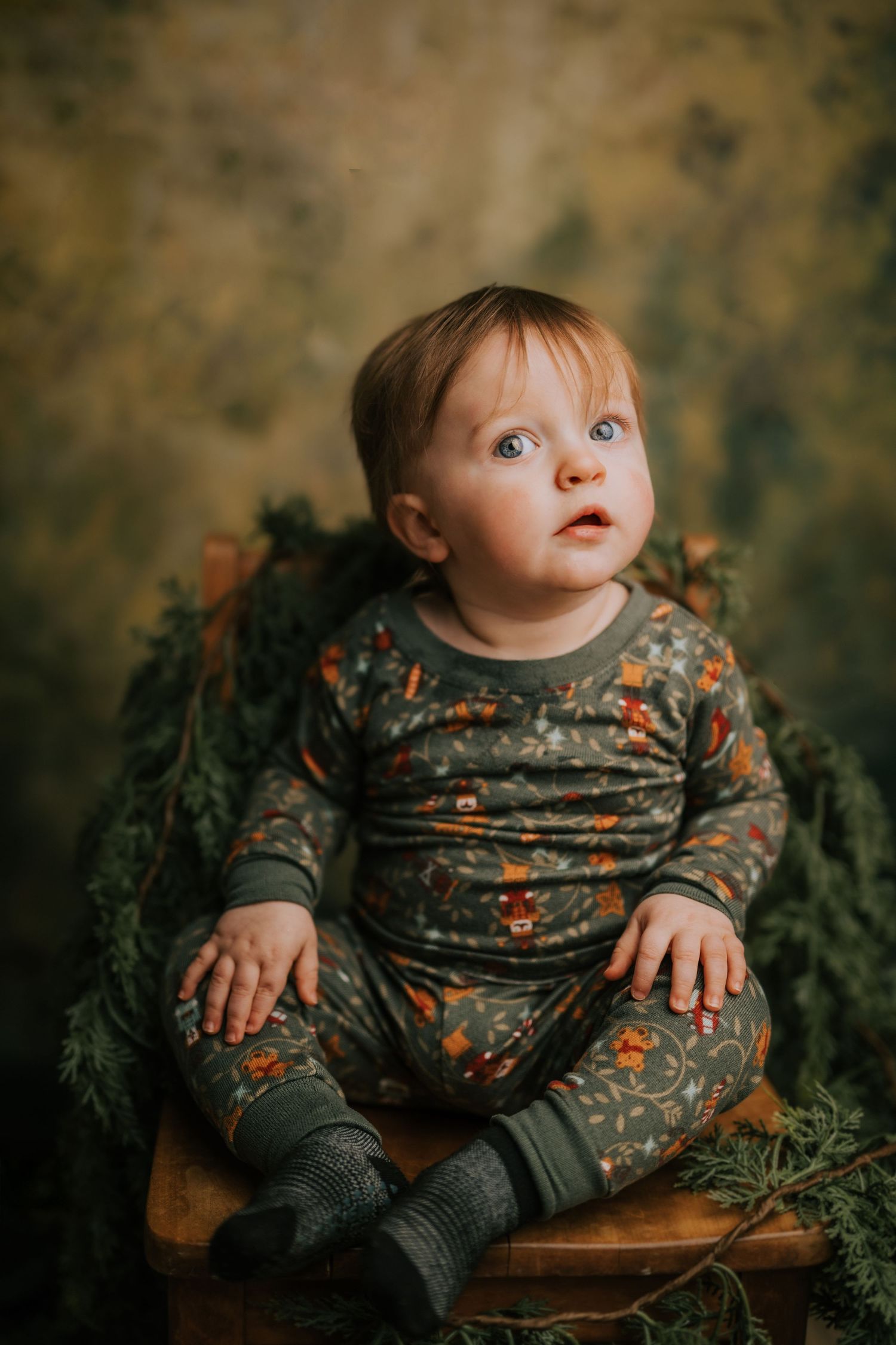 Baby wearing floral print outfit sits in wooden chair surrounded by greenery.