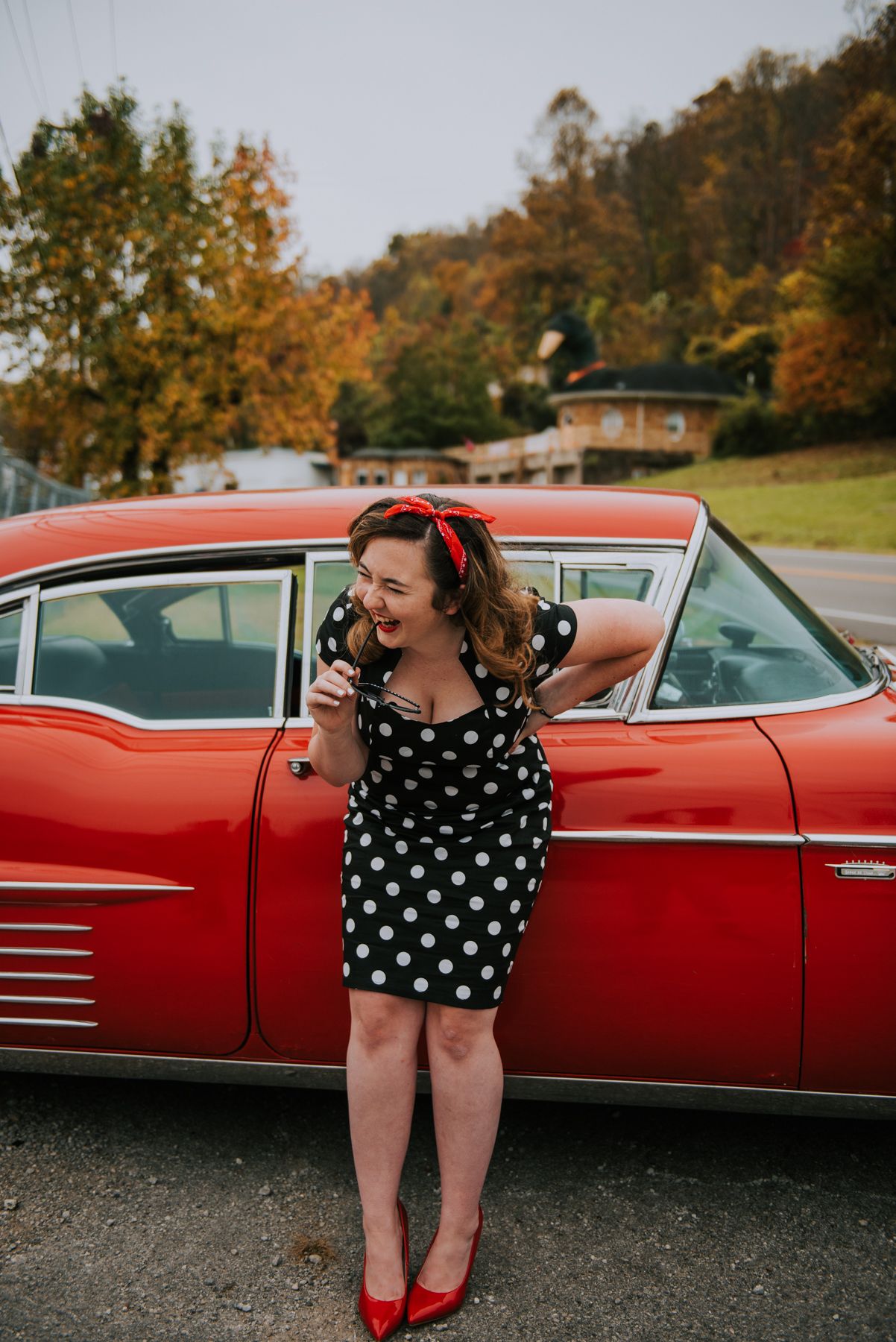 A retro polka dot dress and red heels pose alongside a classic red vintage car.