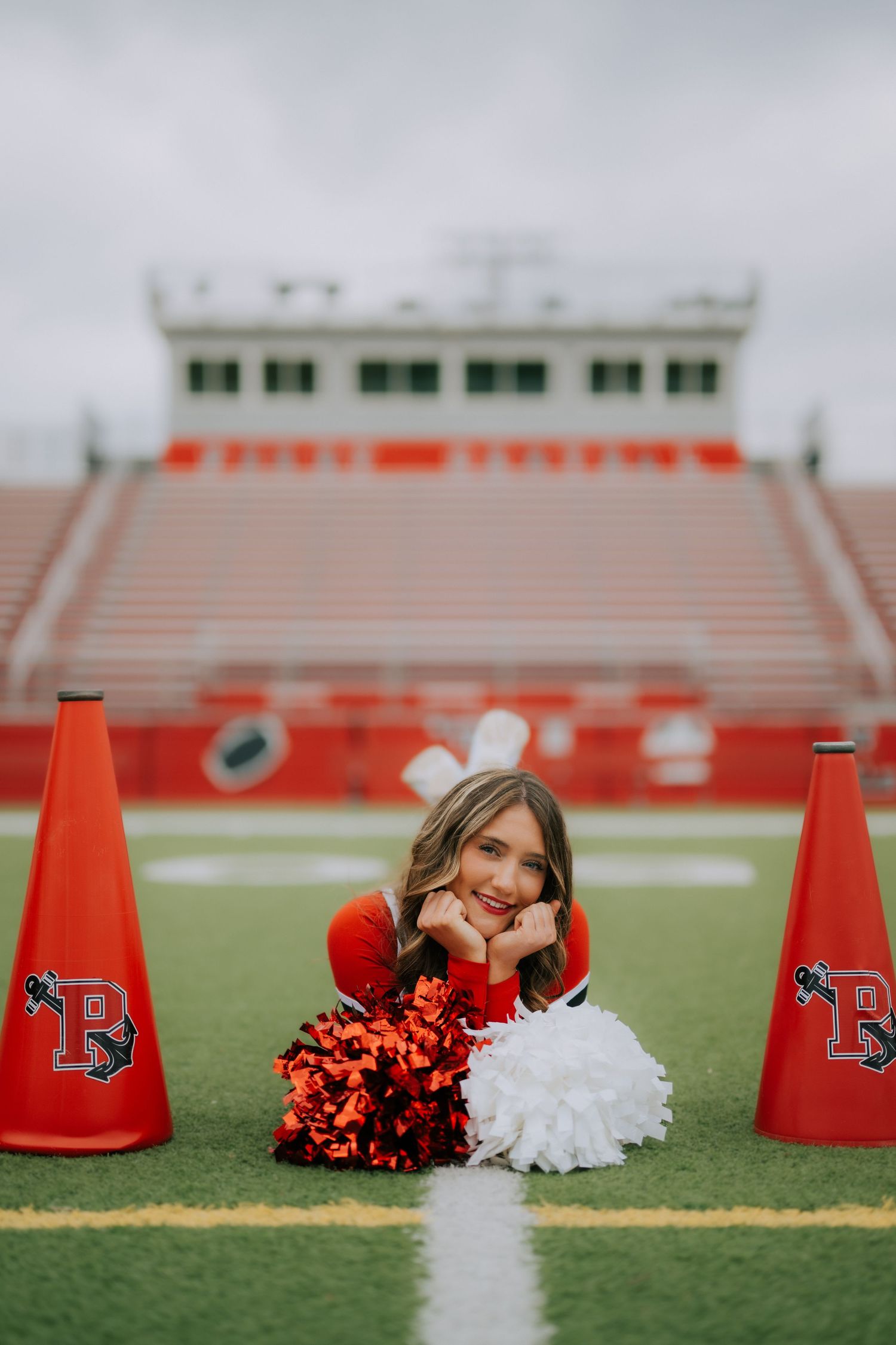 A cheerleader poses with red and white pom poms and megaphones on a football field with stadium bleachers in background.