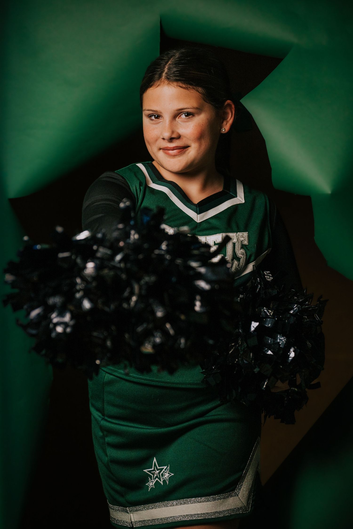 Cheerleader in green uniform with black pom poms poses against dark green background in series of photos.
