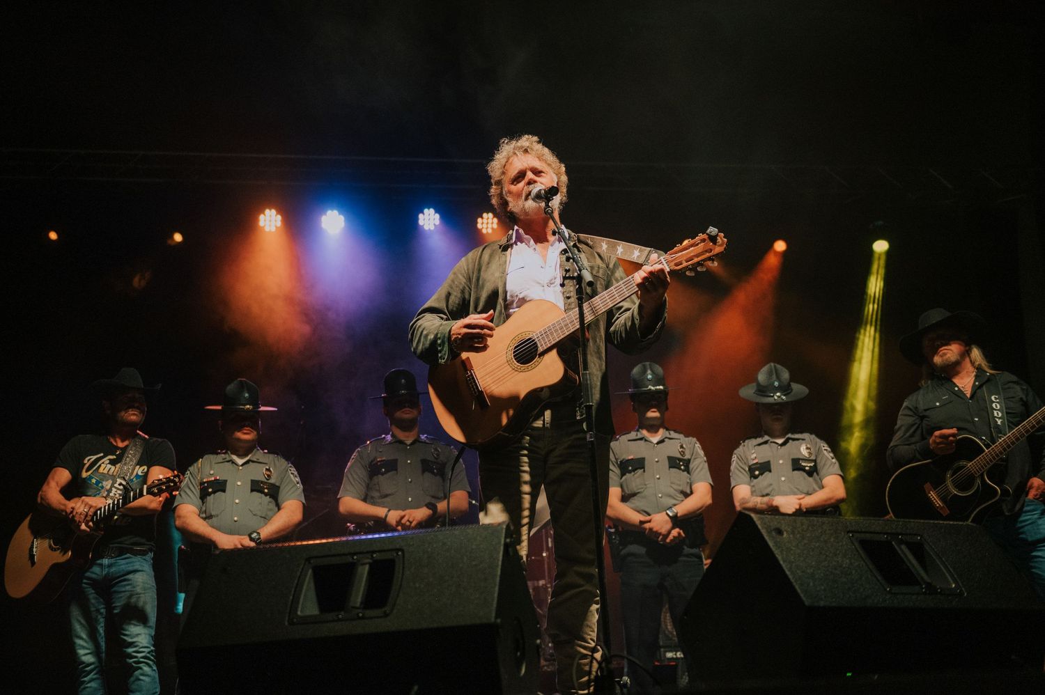 Musician with acoustic guitar performs center stage with band members sitting behind on elevated platforms.