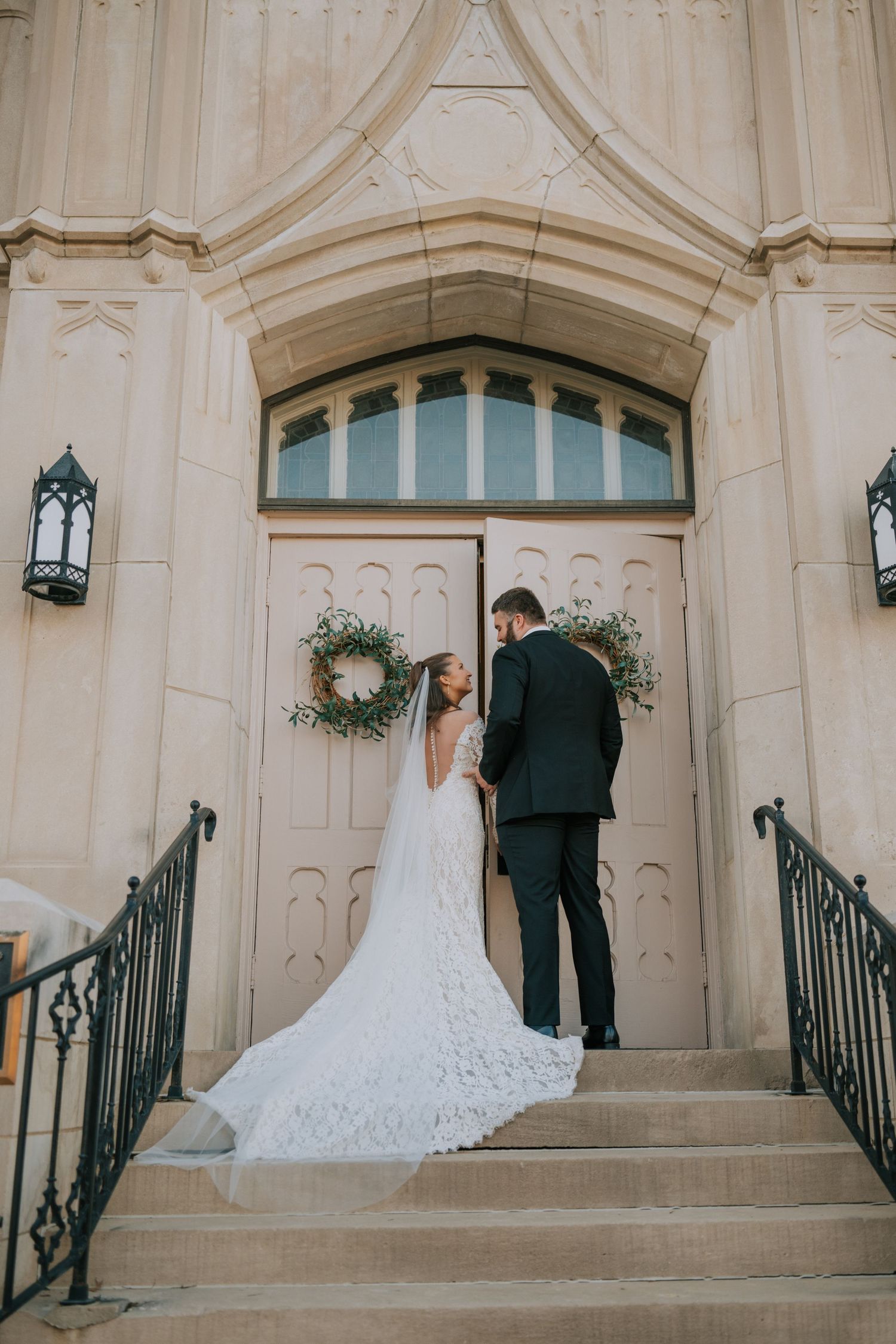 A wedding couple stands at church entrance steps with ornate railings and a wreath-adorned door in cream stonework.