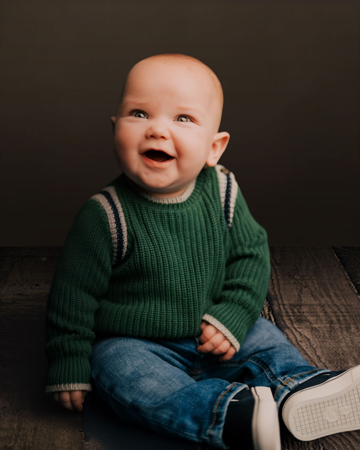 Smiling baby in green knit sweater and jeans sitting cross-legged on wooden floor against dark background.