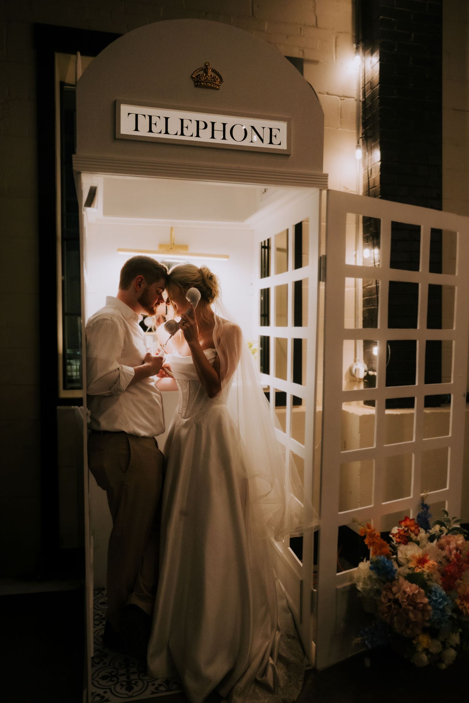 Bride and groom share romantic moment inside white vintage telephone booth at night wedding reception.