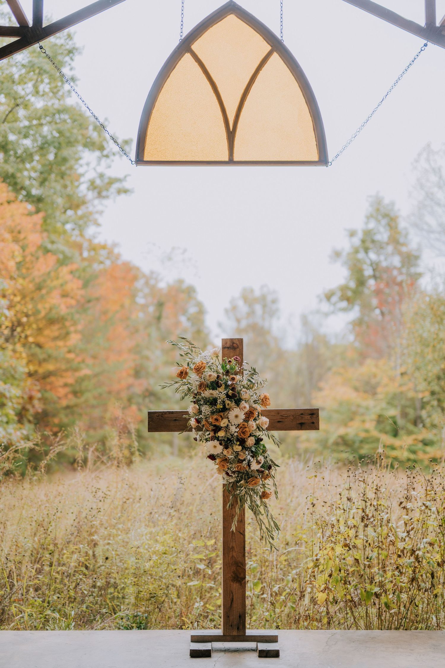 A rustic wooden cross adorned with autumn flowers stands in a field of tall grass with fall foliage in the background.