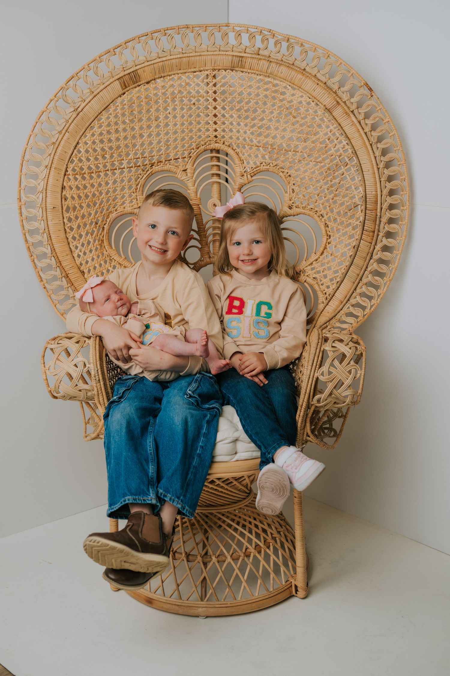 Two young children sit together on a large woven peacock chair against a white wall.