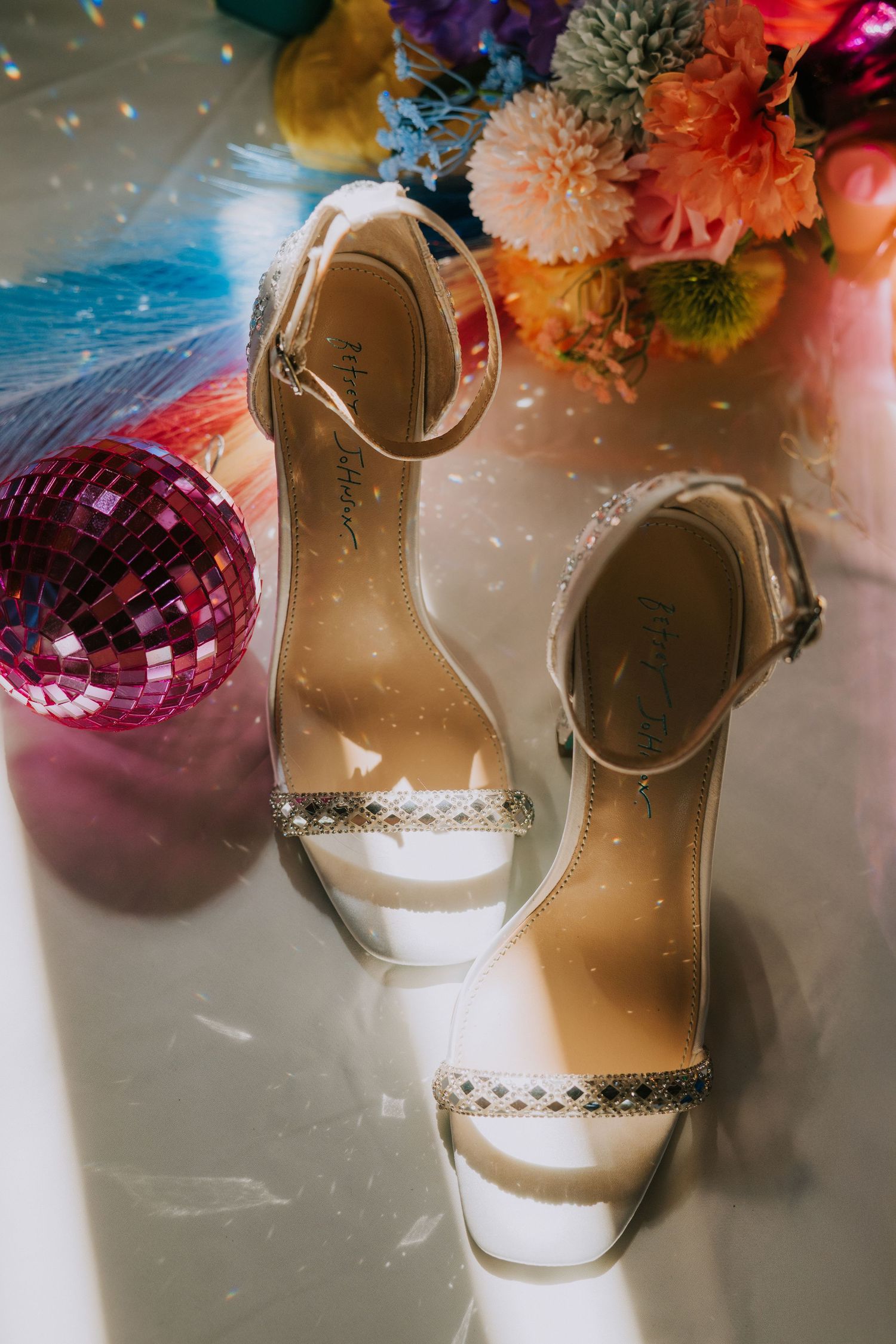 White bridal sandals with rhinestone details next to pink disco balls and colorful flowers.