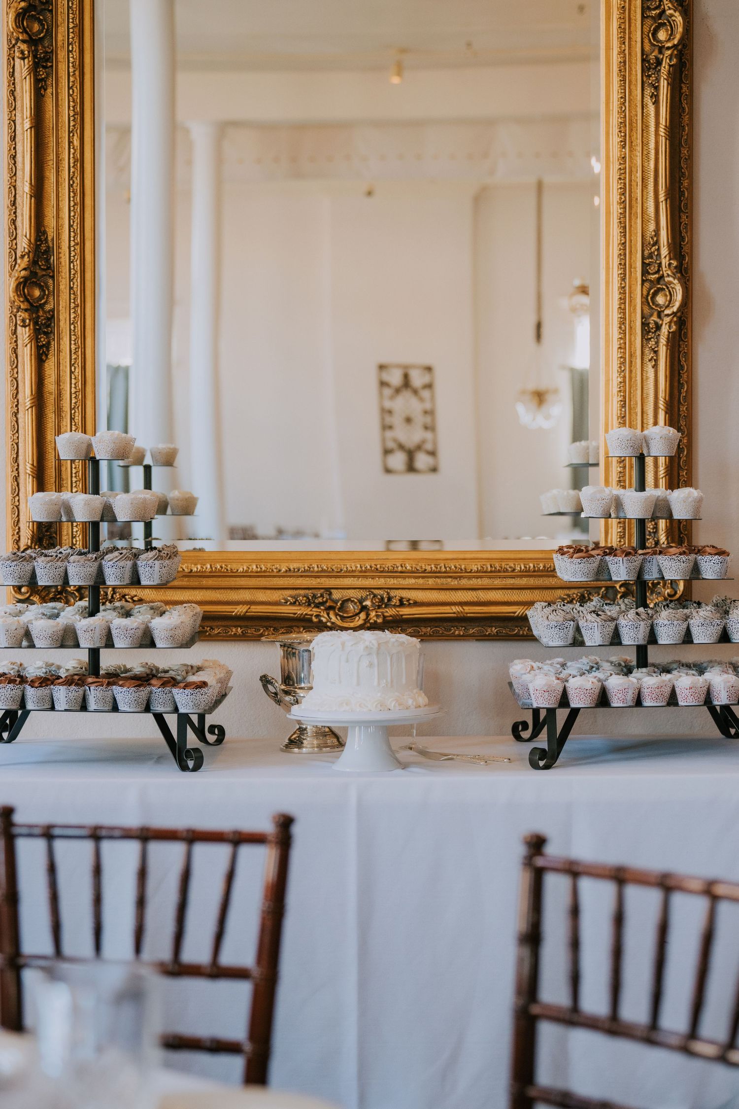 Elegant wedding dessert display with tiered cupcake stands and a white cake centered in front of an ornate gold mirror.