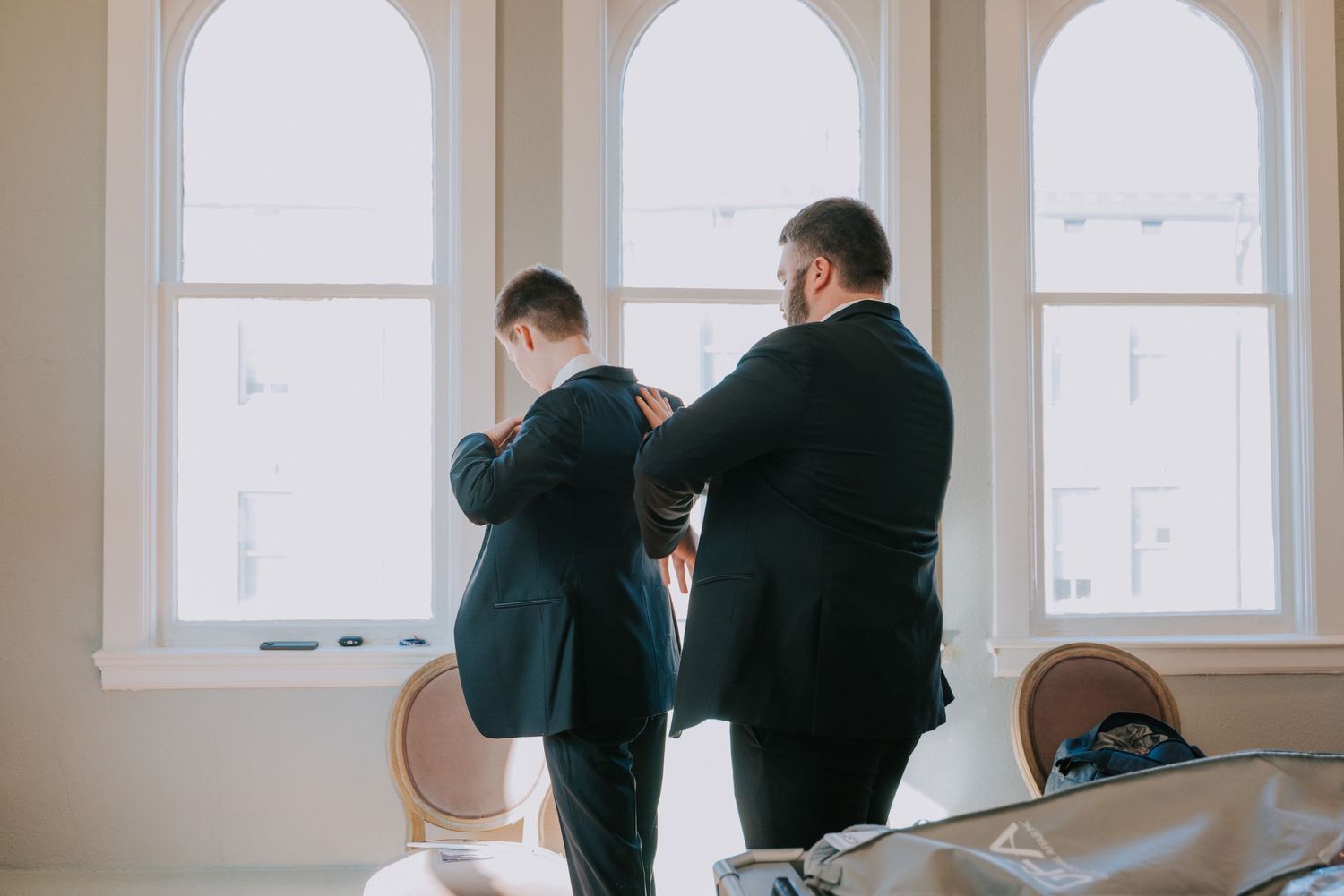 Two people in formal black suits stand by bright arched windows in an elegant room.