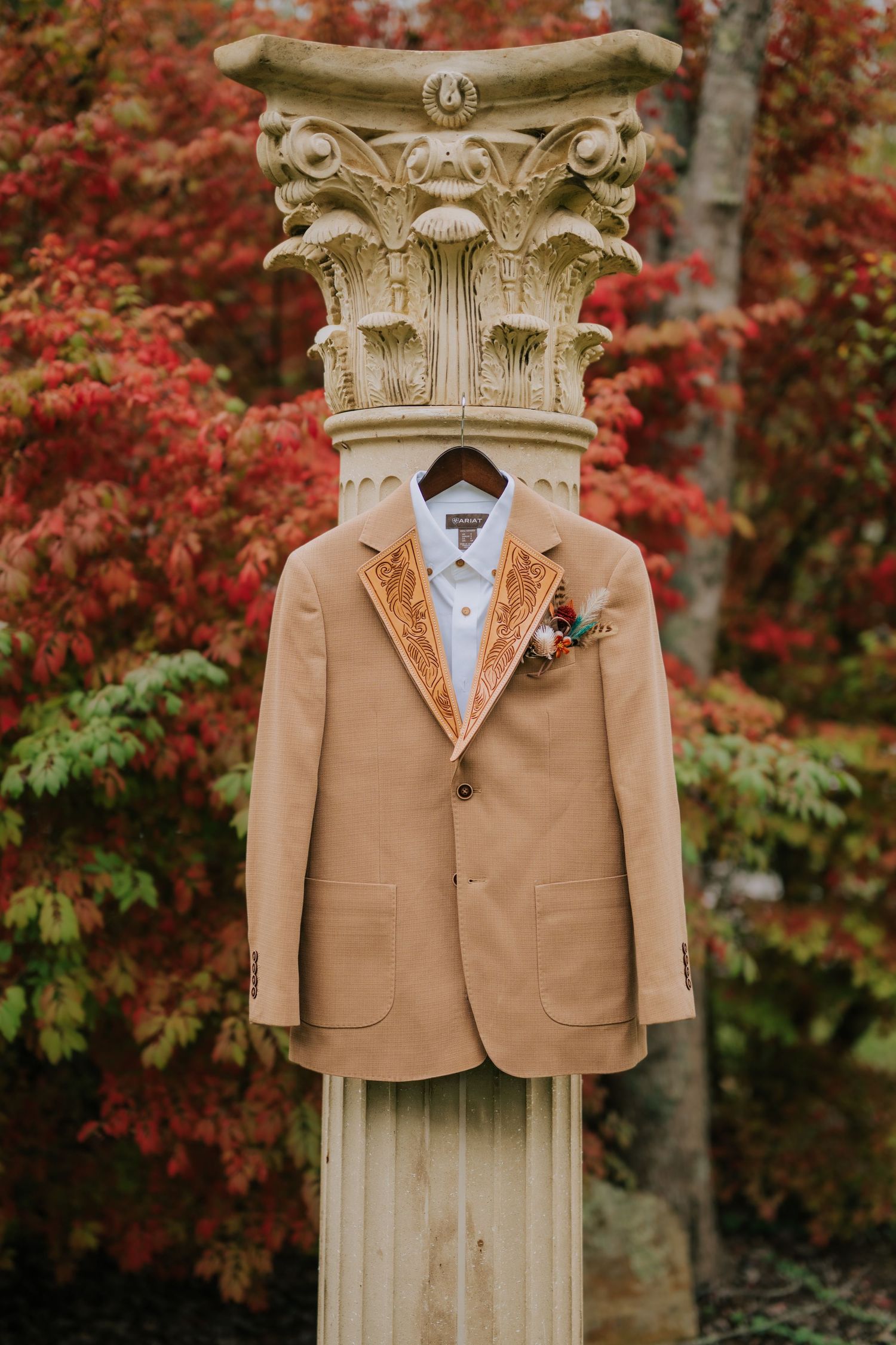 Tan blazer with floral boutonniere on stone column, autumn red foliage backdrop, wedding attire display.