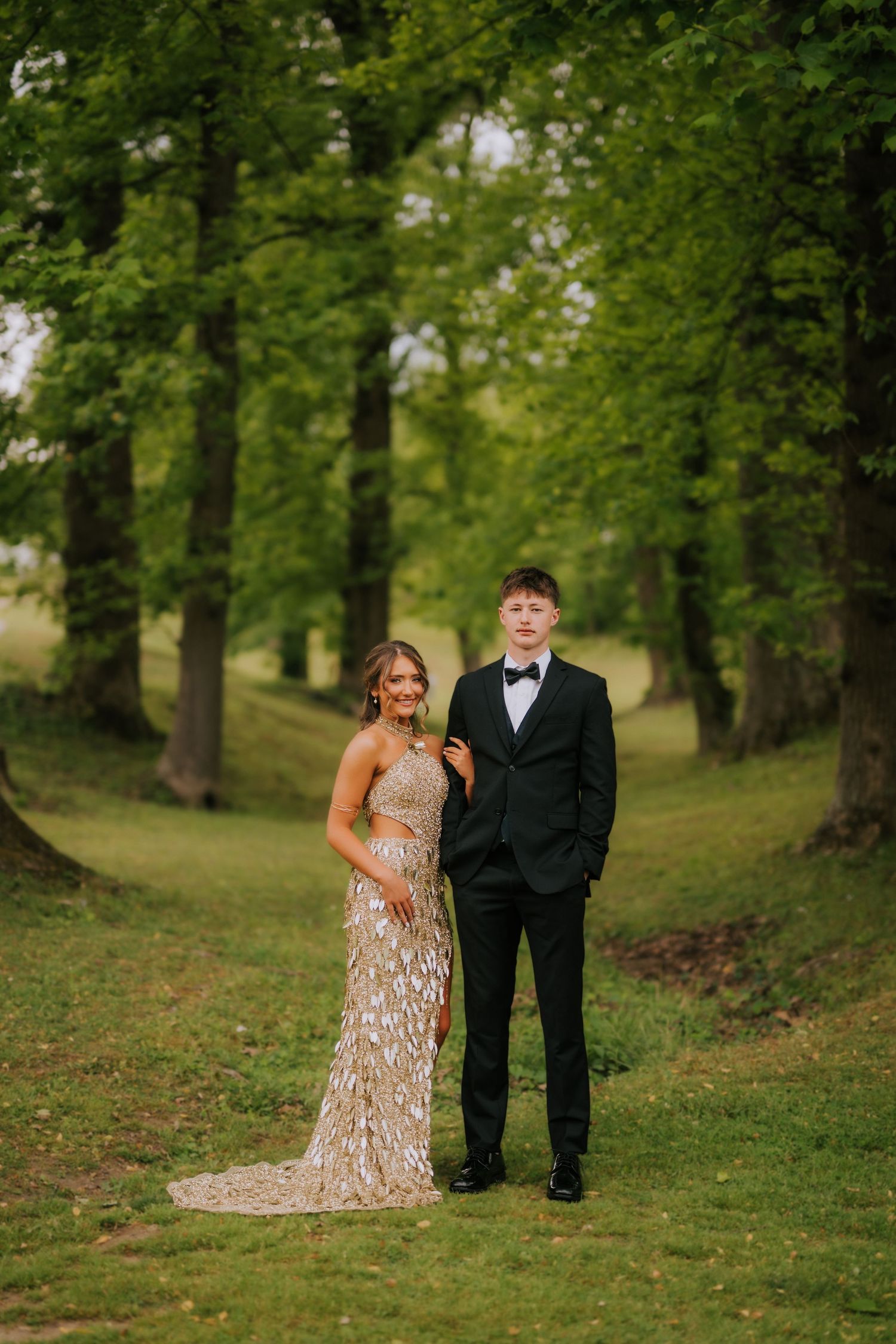 A young couple in formal attire poses outdoors, she in a gold gown and he in a black tuxedo, surrounded by lush green trees.