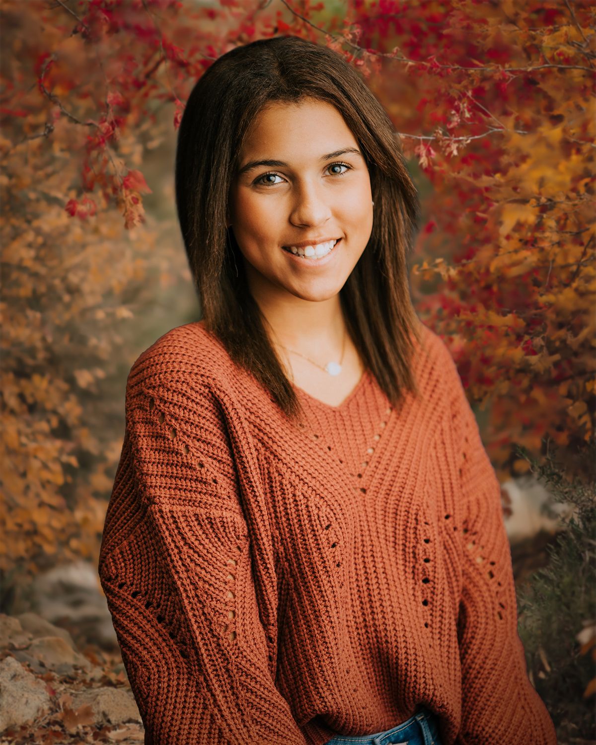 Young woman in rust knit sweater smiling outdoors among vibrant autumn foliage backdrop.