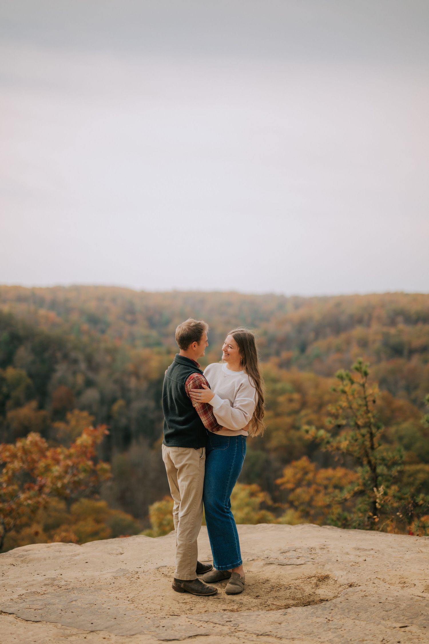 Two people embrace on a rocky overlook with fall colors in the valley below.