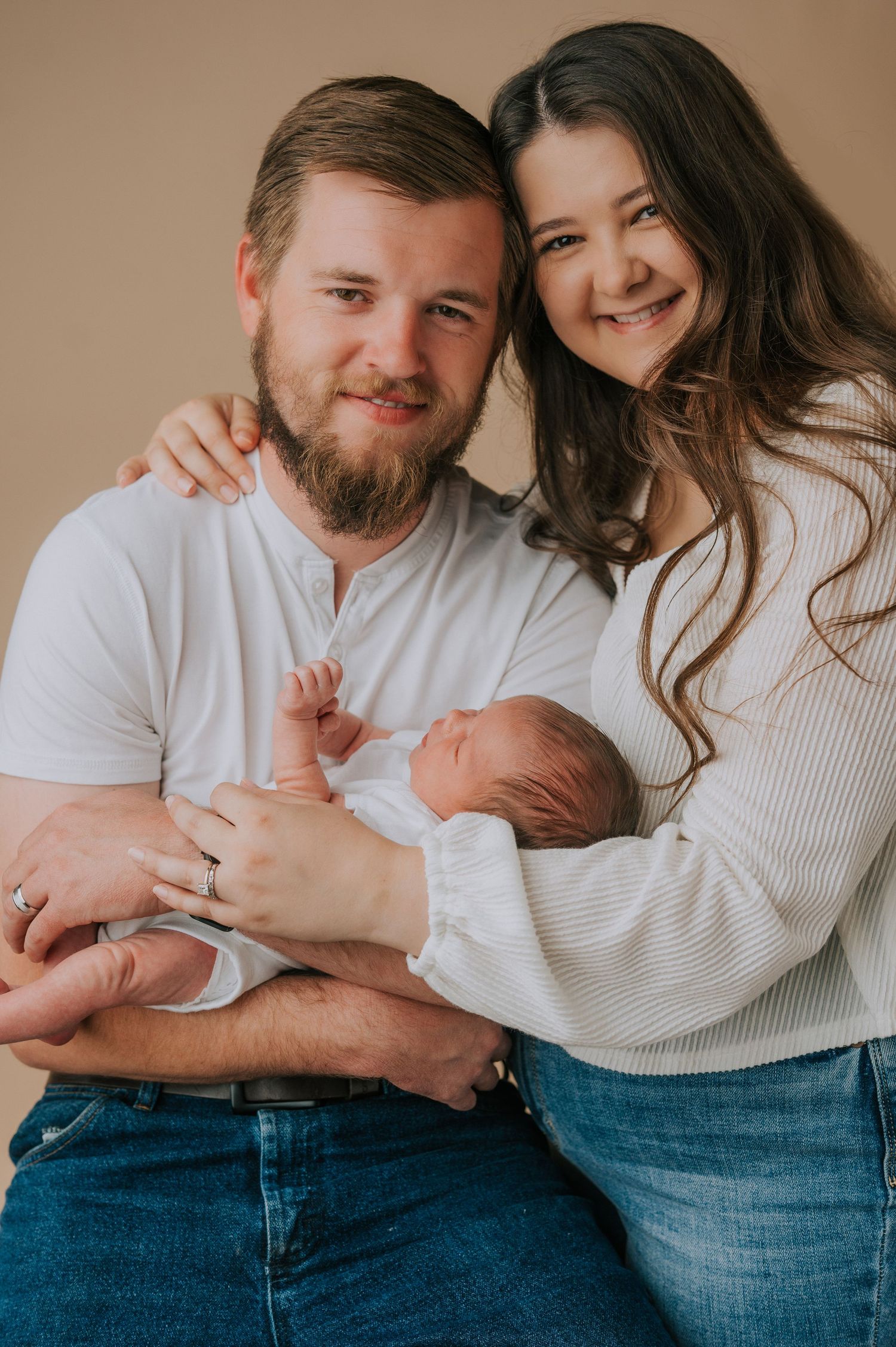 Couple in casual attire holding newborn baby against warm brown studio backdrop.