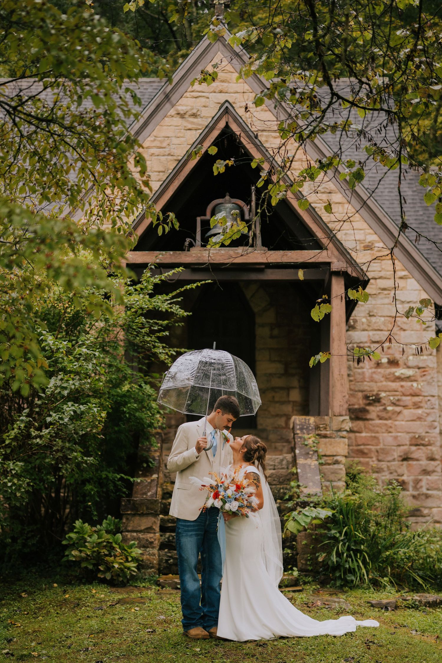 A couple shares a romantic moment under a clear umbrella outside a charming stone chapel.
