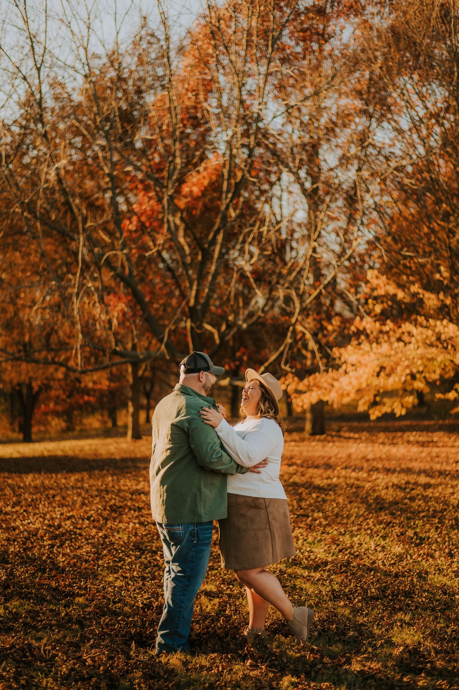 Couple embracing in a field during golden hour with autumn foliage in the background.