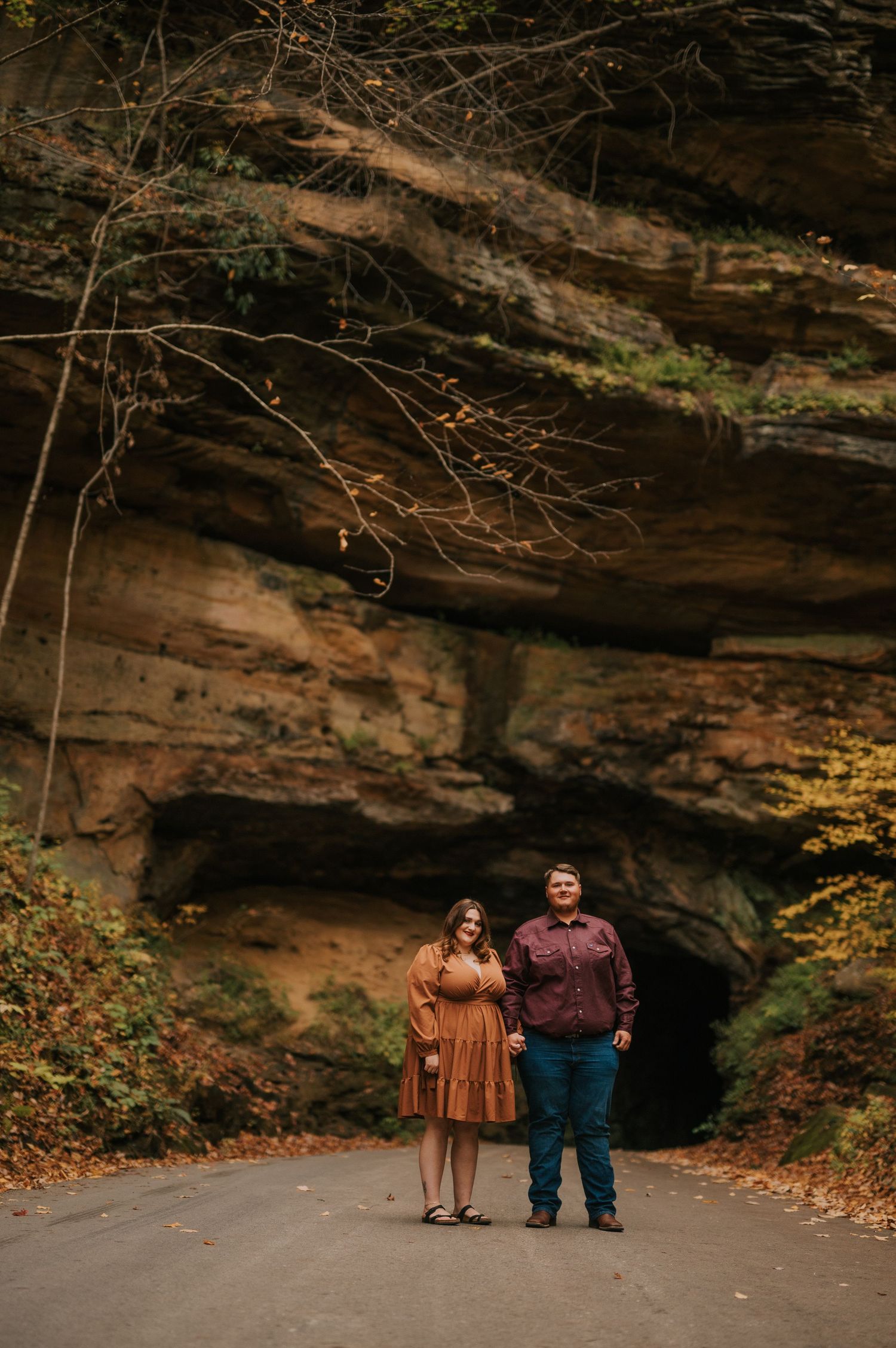 A couple stands together on a path beneath a dramatic rocky cave entrance surrounded by autumn trees.