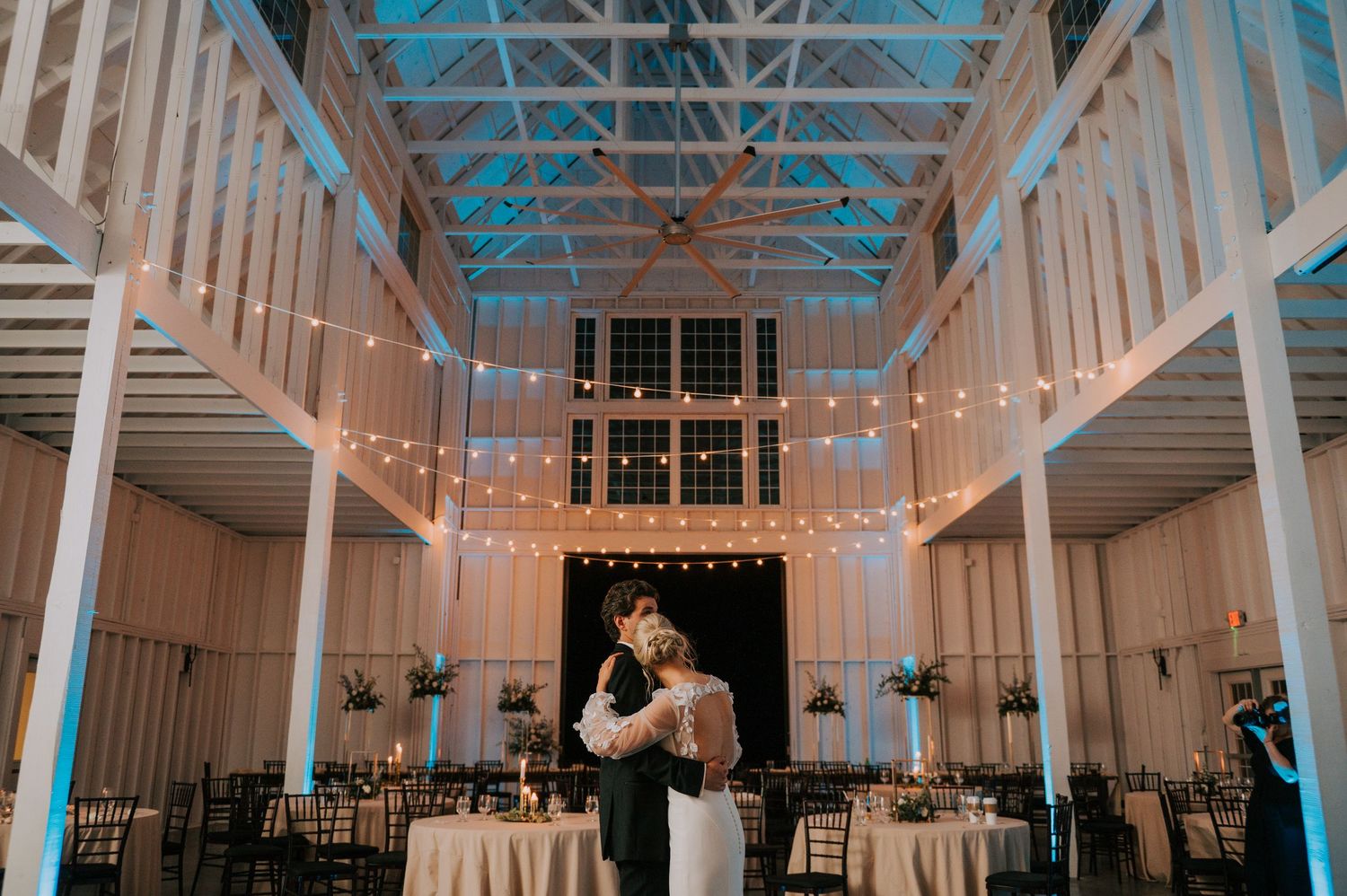 Reception venue with high ceilings decorated with string lights and white draping.