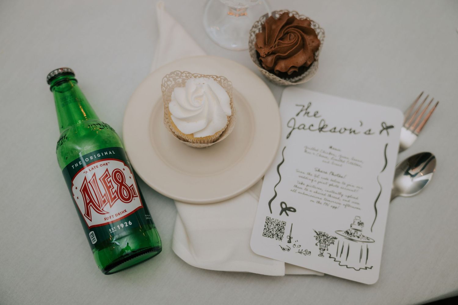 Wedding reception details with Ale-8 bottle, cupcake, and handwritten menu card on white surface.