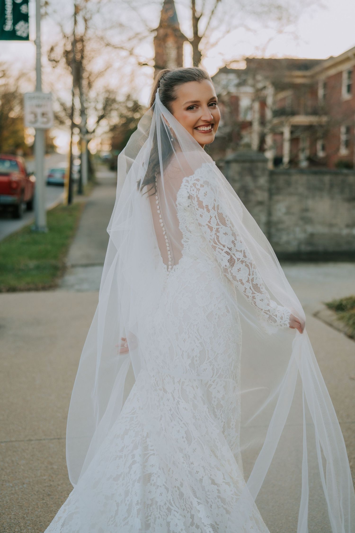A stunning bride in a lace wedding dress with long sleeves and flowing veil poses on a sidewalk.