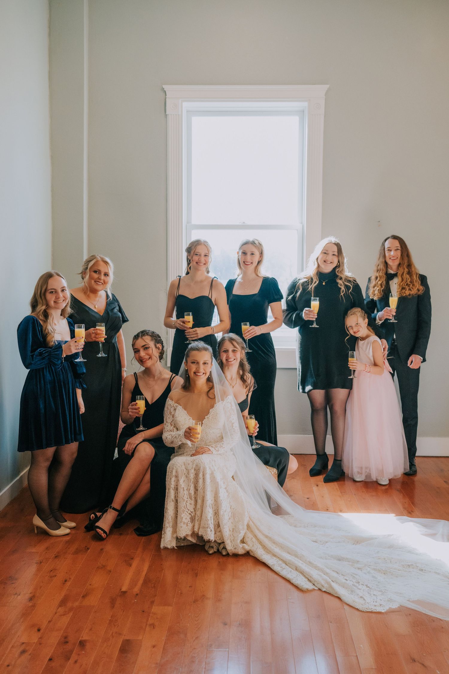 A bridal party in navy blue dresses gathers for a candid photo with the bride on hardwood floors.