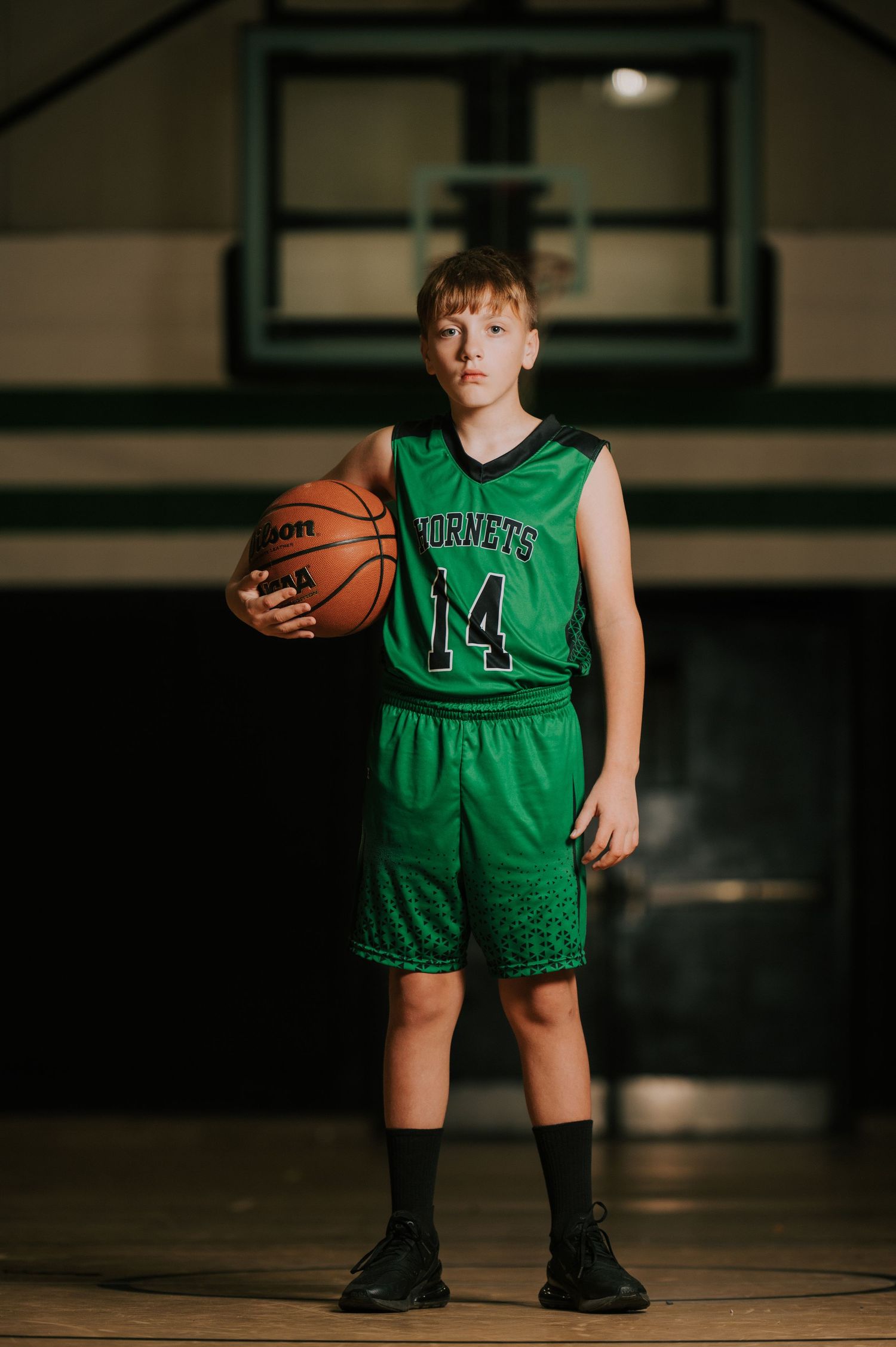 A young basketball player in a green Hornets jersey stands on a court holding a basketball in dramatic lighting.