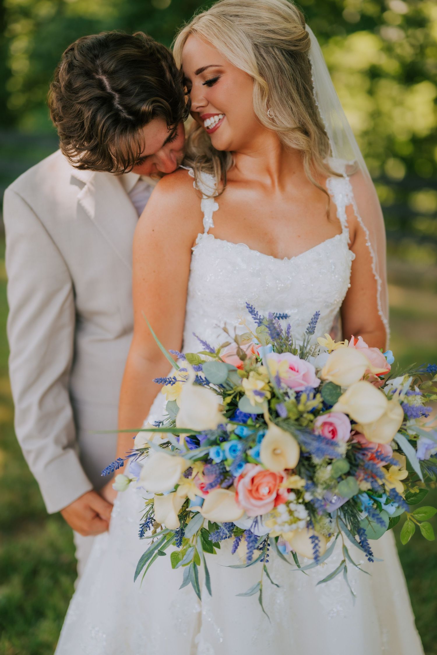 Bride in lace gown holds colorful bouquet with roses and lavender while groom in tan suit nuzzles her cheek outdoors.