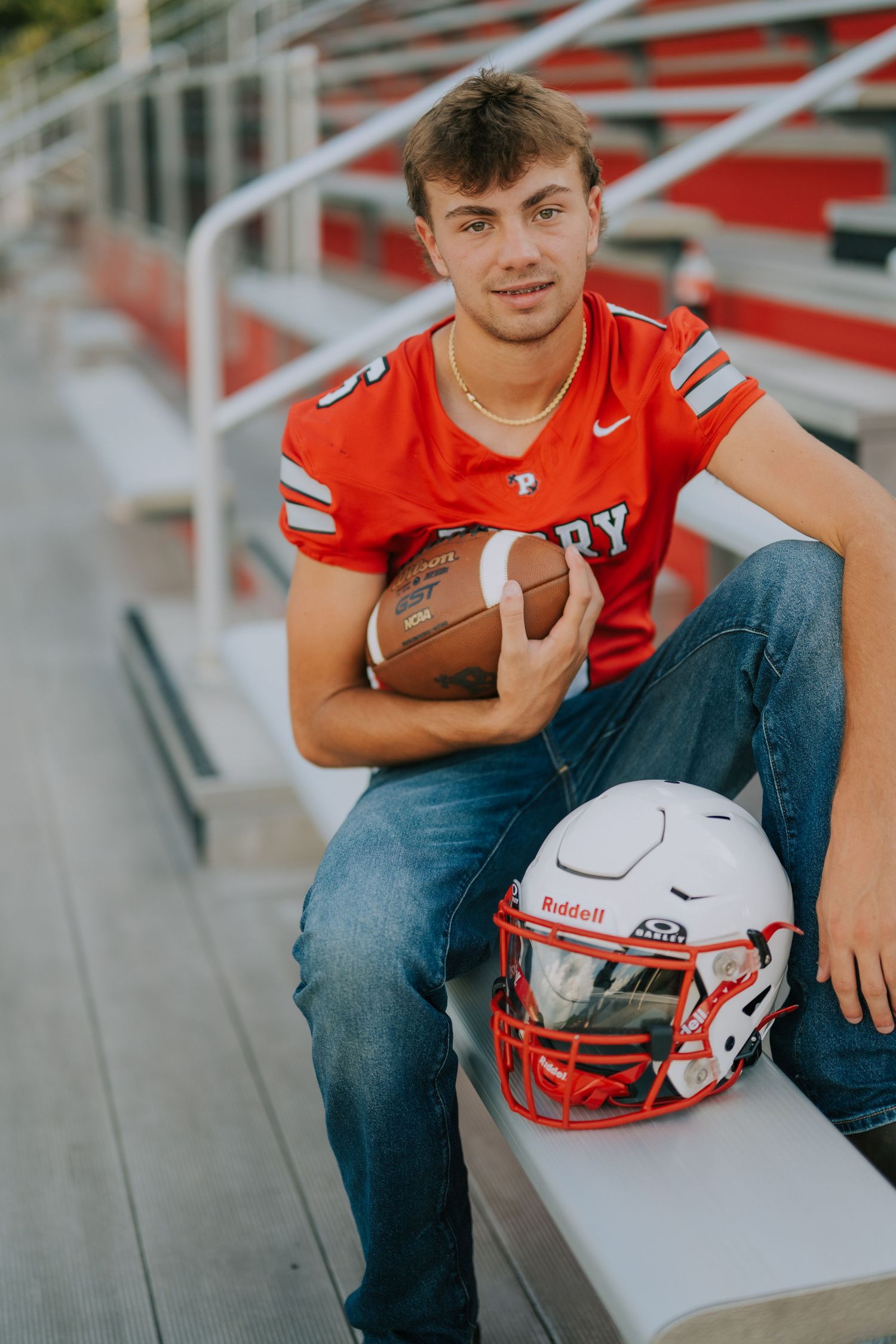 A football player in a red jersey sits on bleachers holding a ball and helmet.