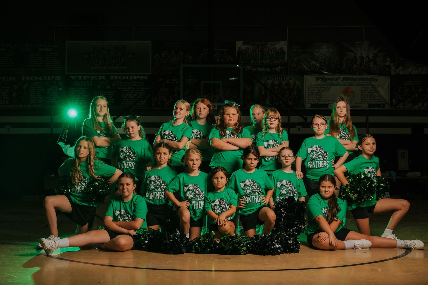 Cheerleading team in green uniforms poses together for group photo on gymnasium floor.