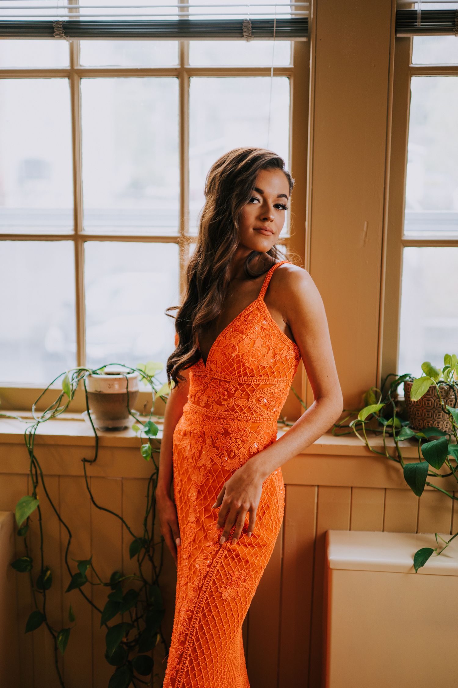 Fashion portraits by a window featuring an orange lace dress with natural lighting and indoor plants.