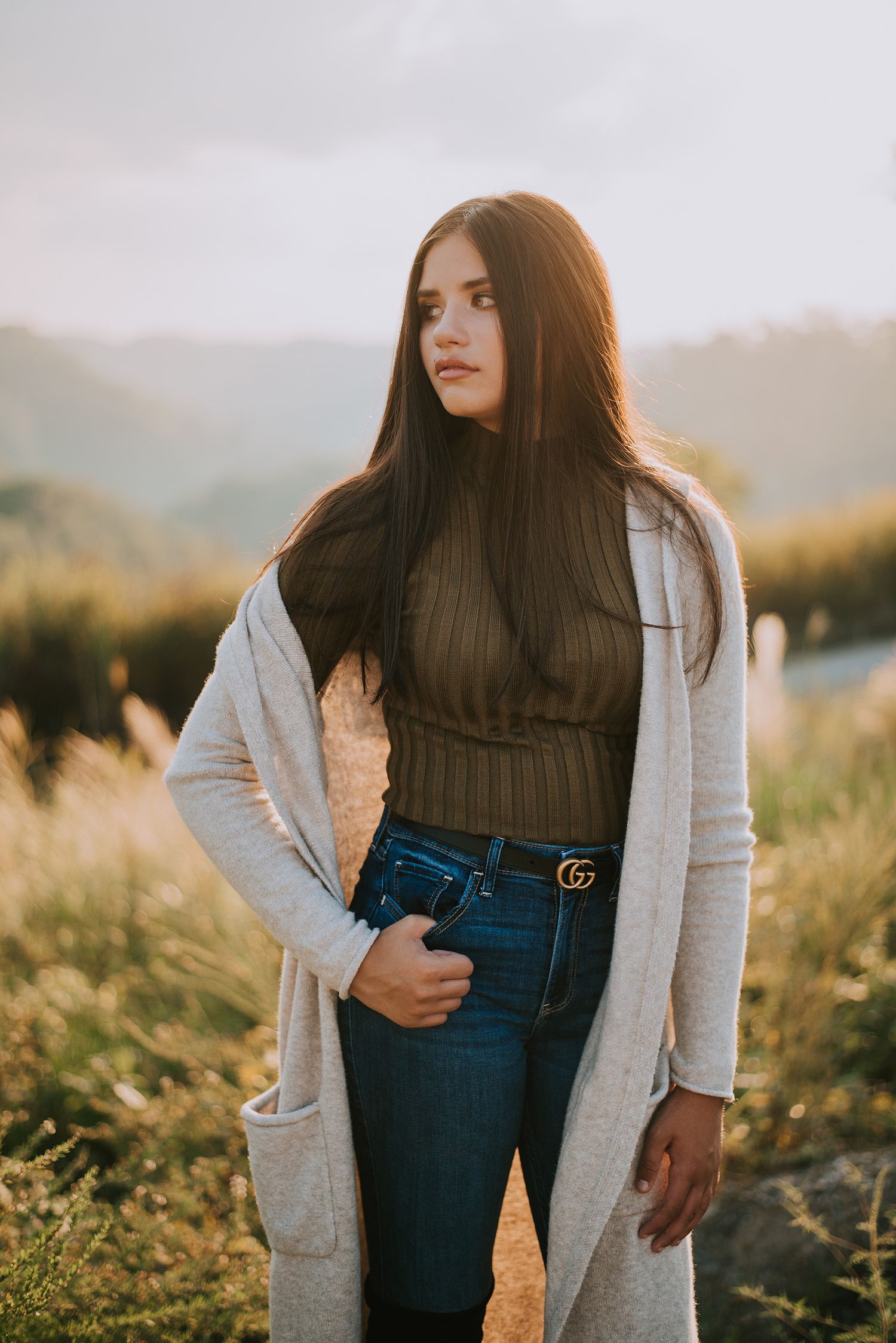 A fashionable fall outfit featuring a black turtleneck, long gray cardigan and designer belt against a golden sunset.