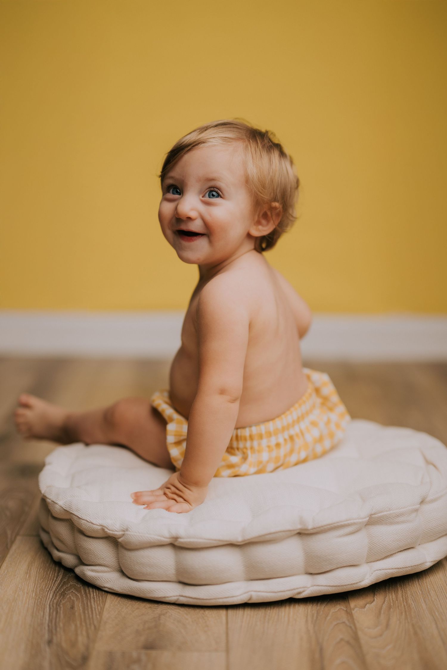 Baby in yellow checkered outfit crawls on tufted cushion against warm yellow background.