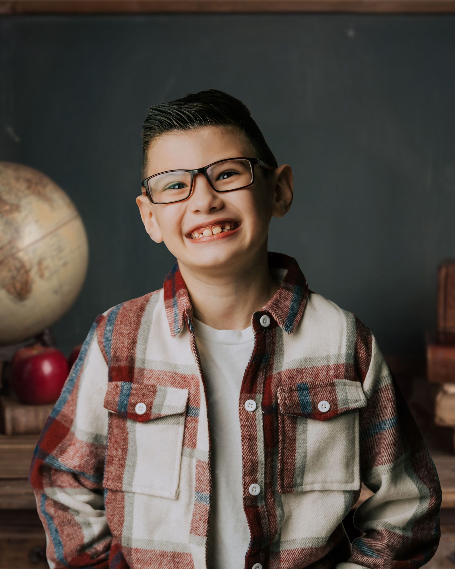 Smiling boy with glasses wearing plaid shirt poses in front of chalkboard with globe and apple in school-themed photo.