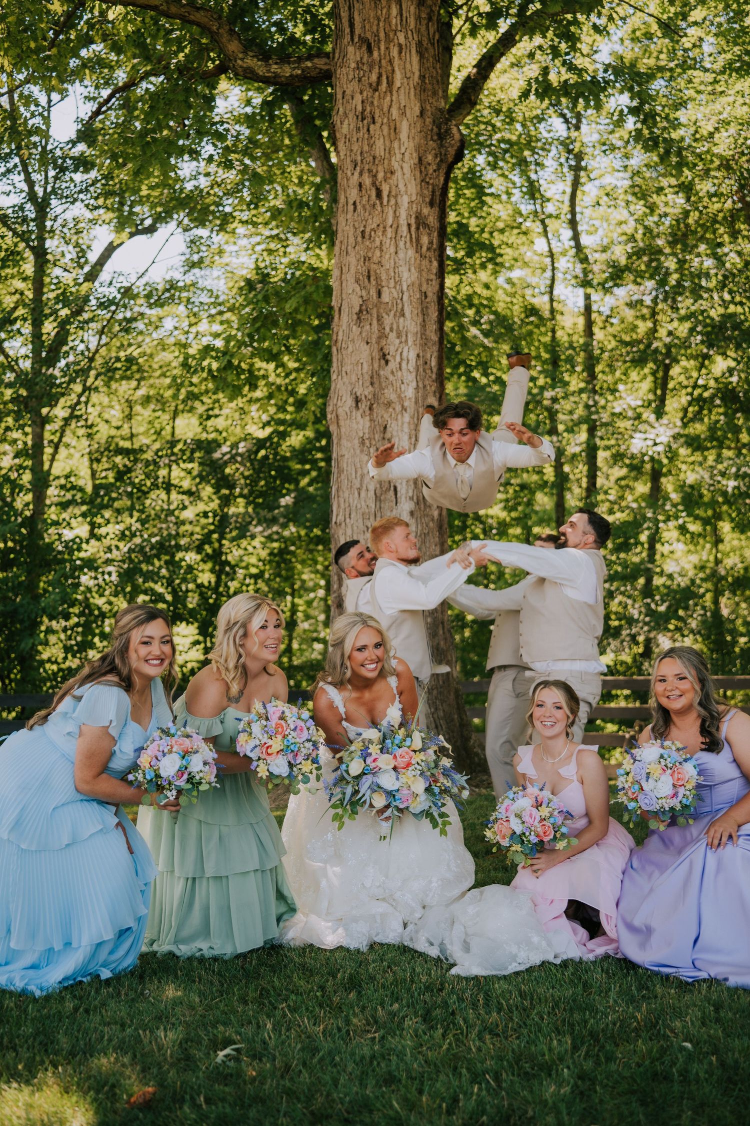 Wedding party poses by a tree, bridesmaids in pastel gowns holding colorful bouquets while groomsmen celebrate behind them.