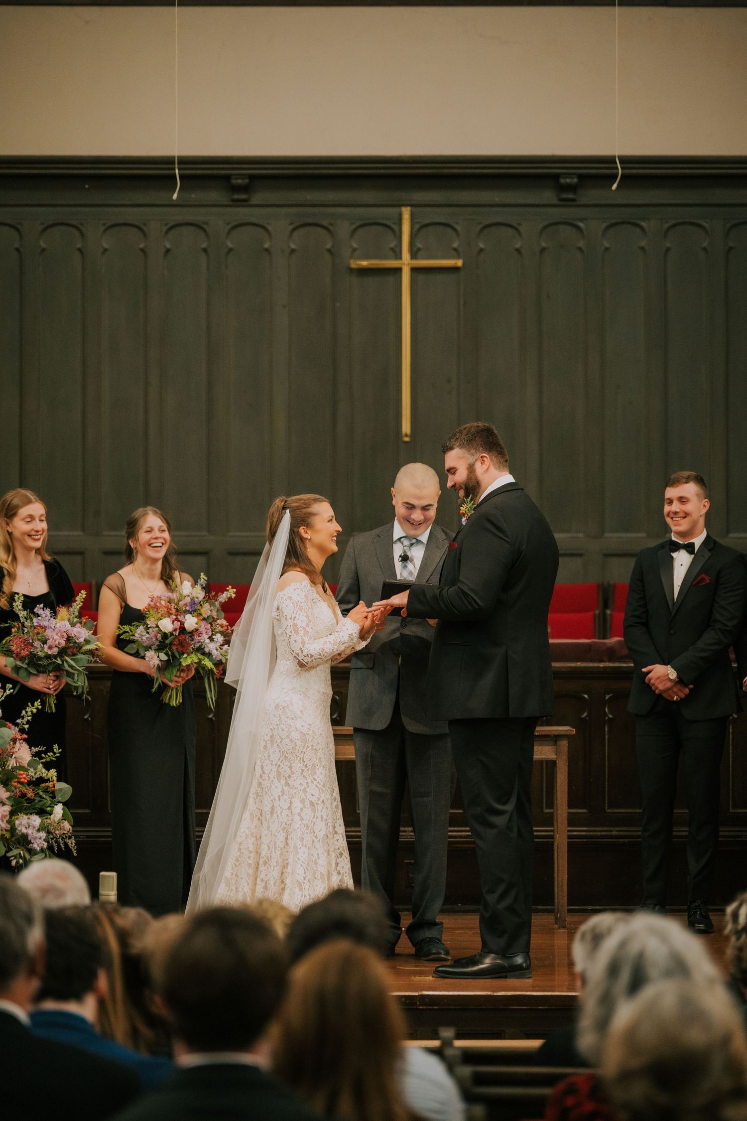 Wedding ceremony taking place at the altar of a church with bridesmaids in black dresses and groomsmen looking on.