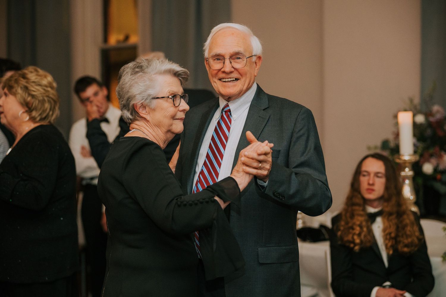 Two people dancing together at an elegant indoor event with other guests visible in the background.