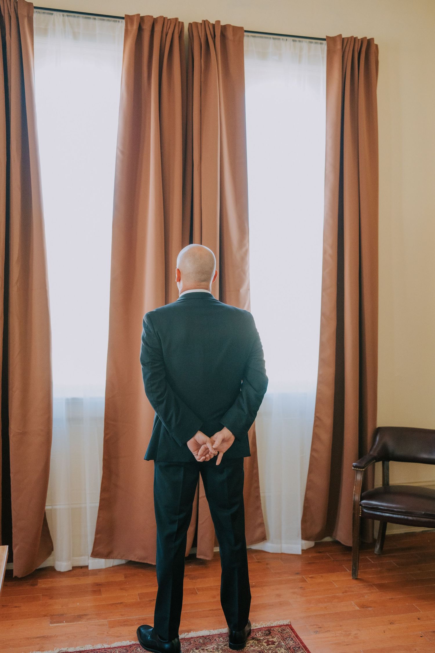 A person in a dark suit stands facing brown curtains with their hands clasped behind their back.