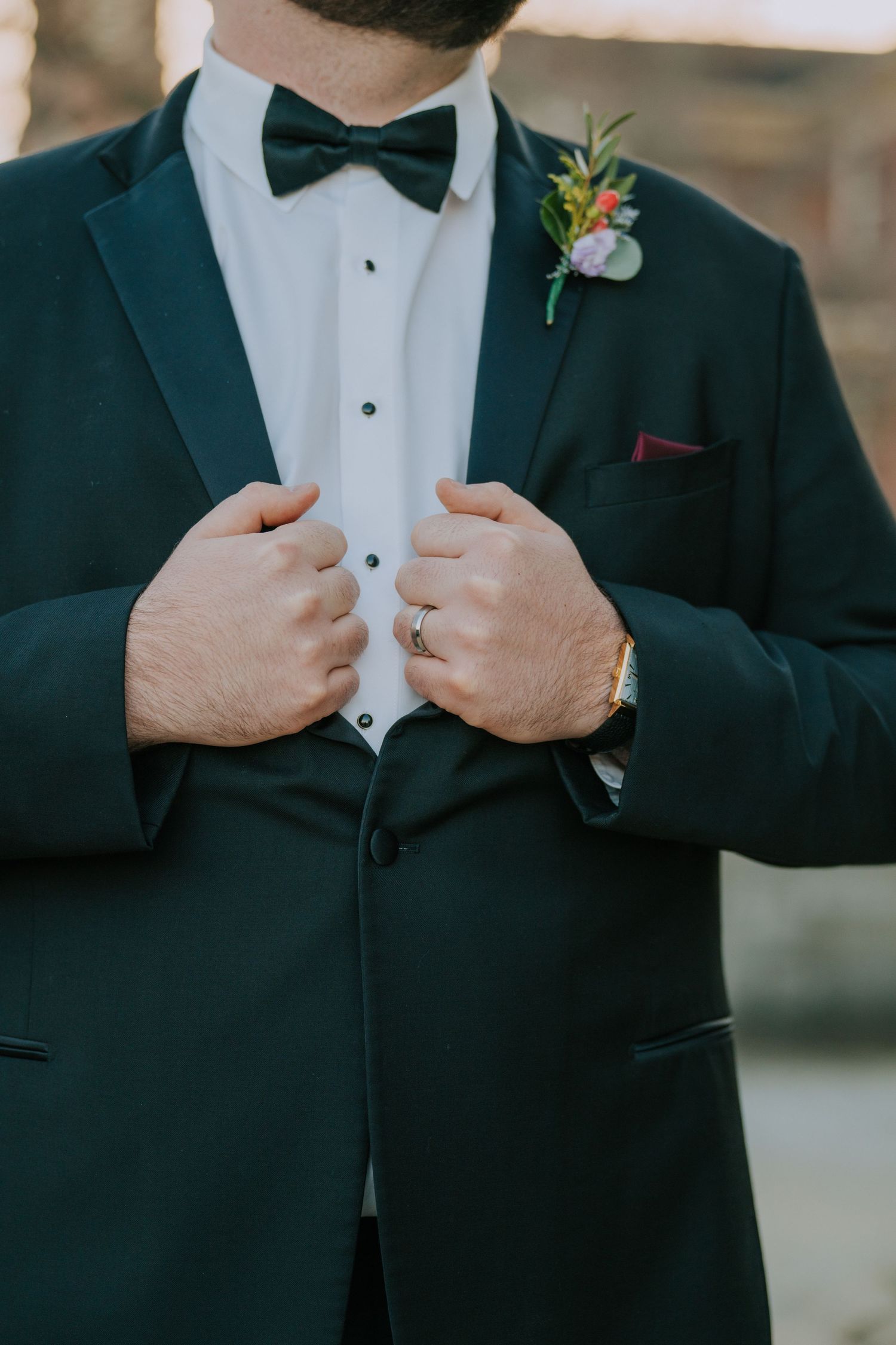 Detail shot of a groom in a dark tuxedo with bow tie and floral boutonniere adjusting his jacket.