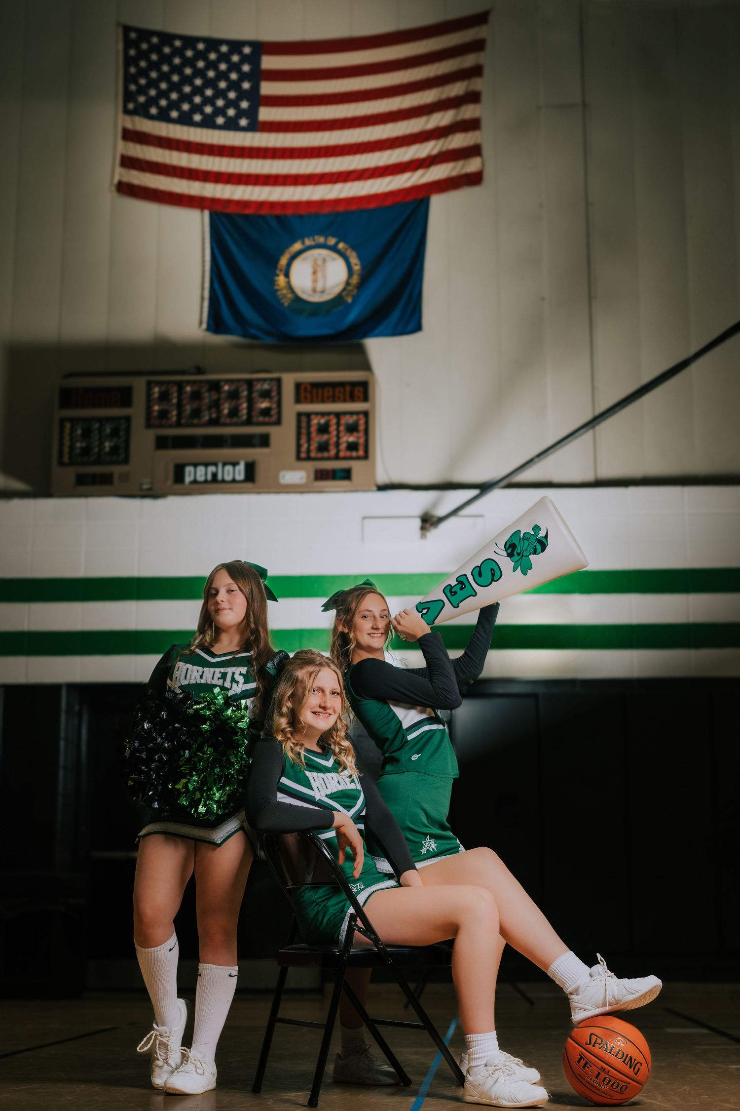 Cheerleader in green uniform poses with basketball in school gym with American flags on wall in dramatic lighting.