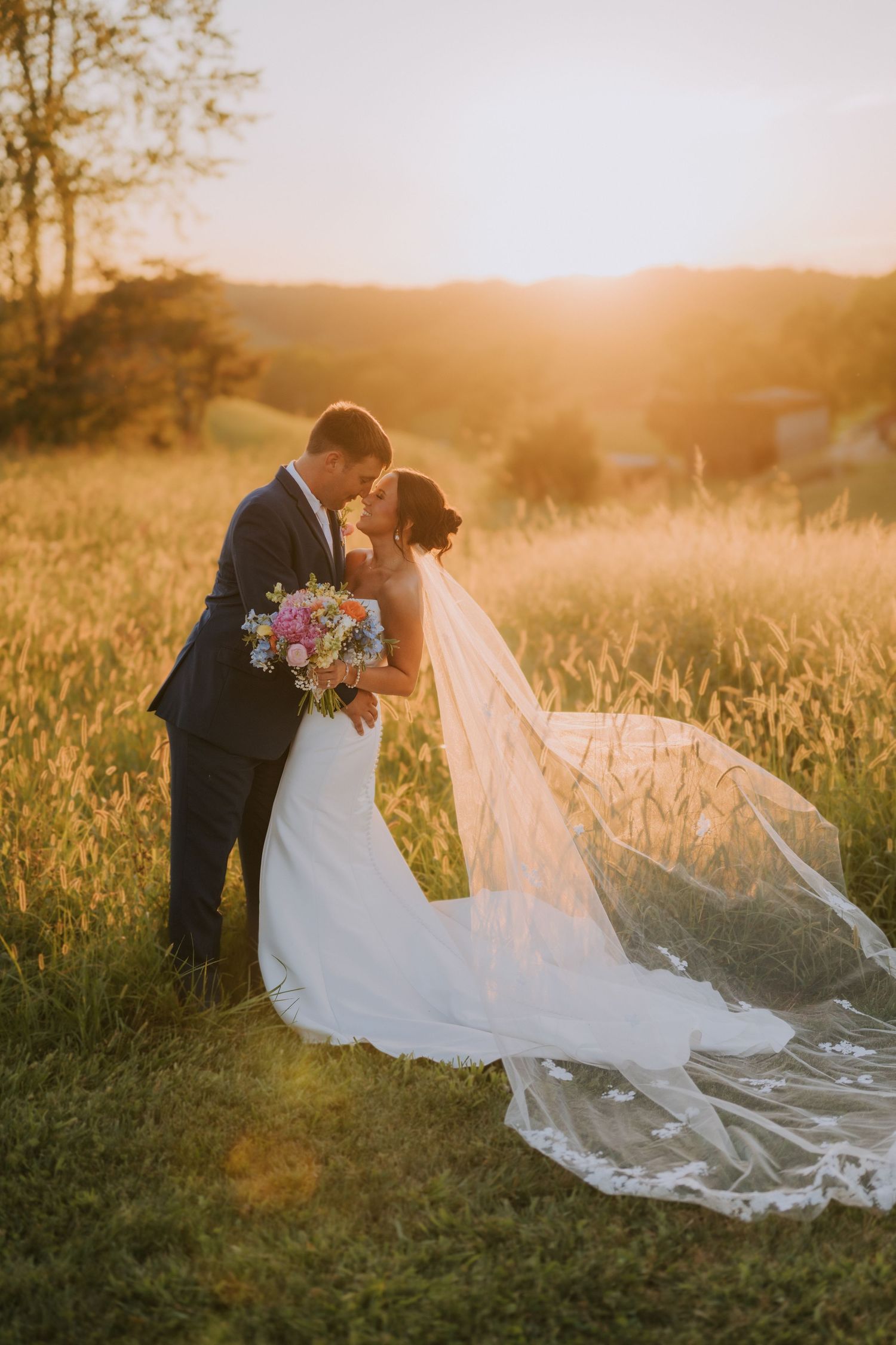 A romantic sunset wedding photo in a golden field with flowing white veil catching the light.