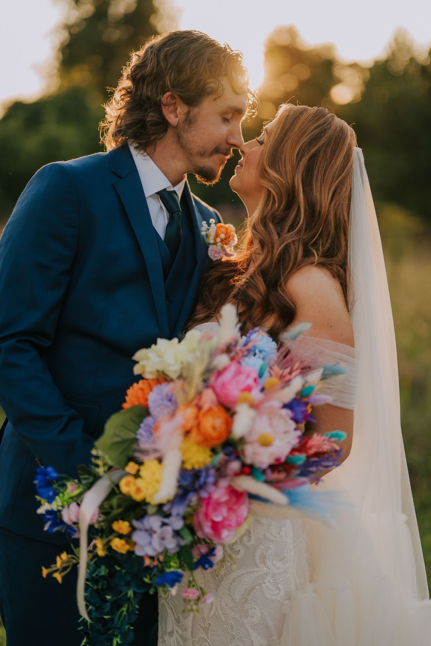A romantic wedding couple embraces in a sunset field with a vibrant multicolored bouquet of flowers.
