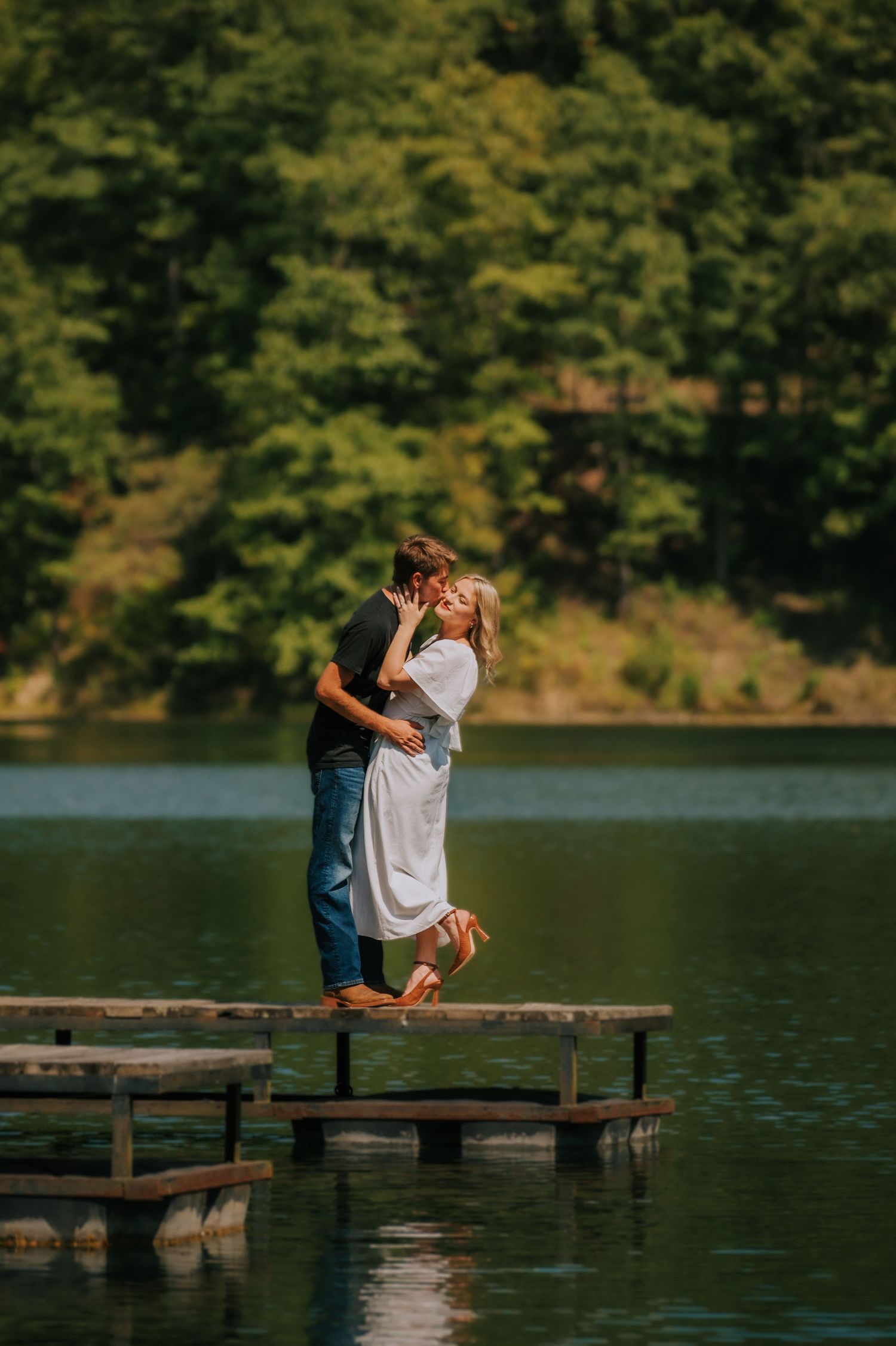 A romantic moment on a wooden dock overlooking a peaceful lake surrounded by green forest.