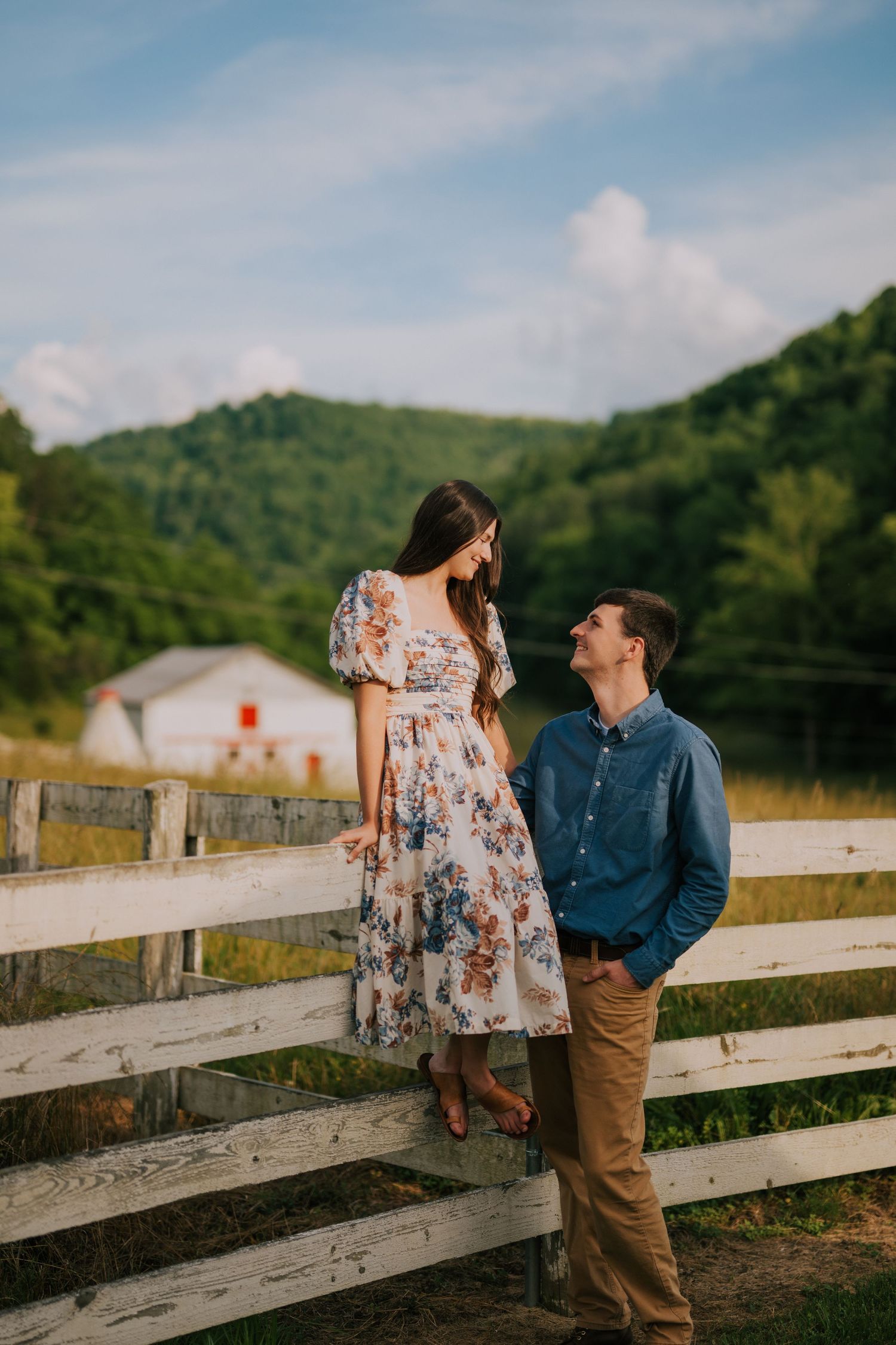 Couple sharing romantic moments by a rustic white fence with rolling hills in background.