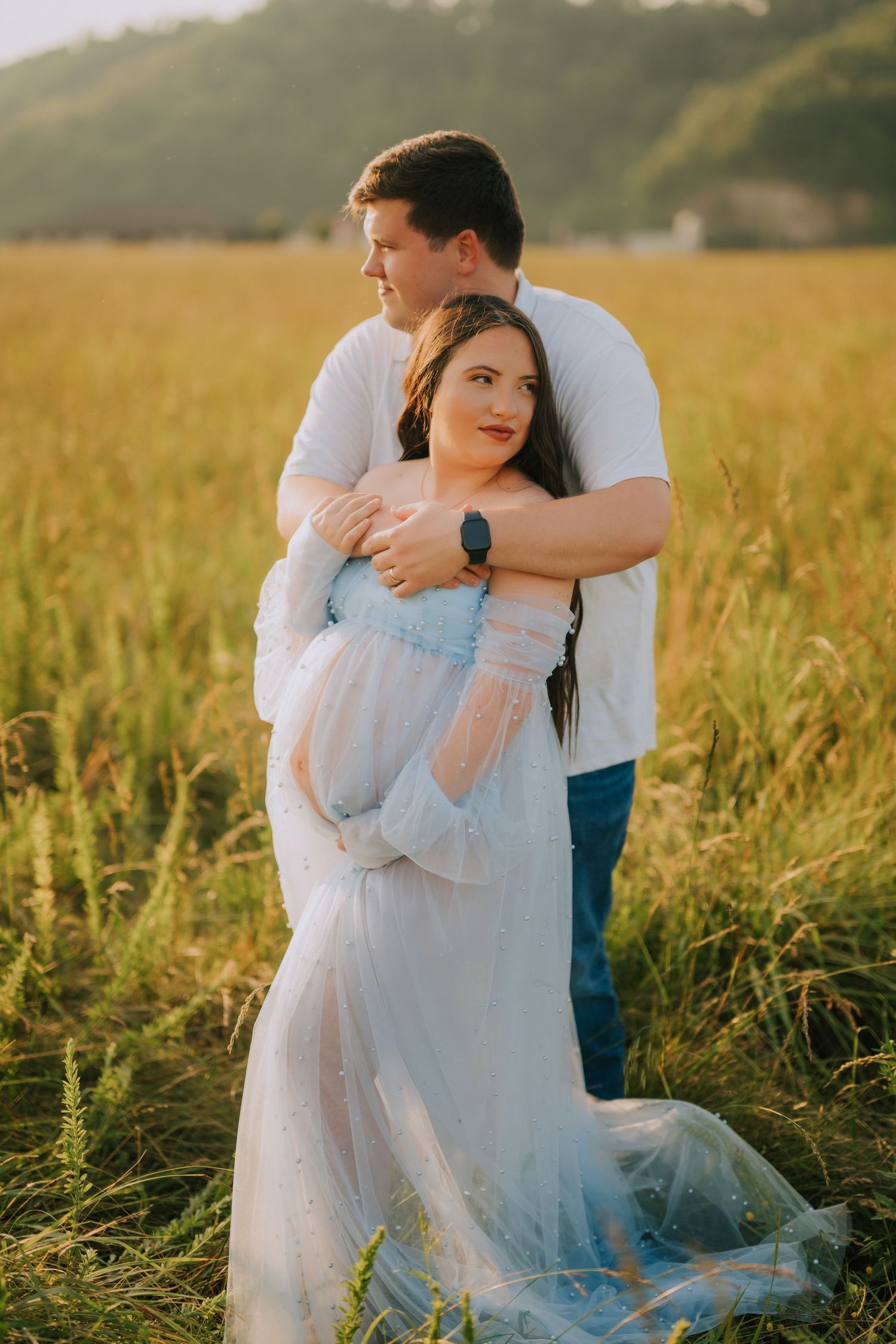 Romantic maternity photoshoot in a sunlit field with couple in light blue dress and casual wear.