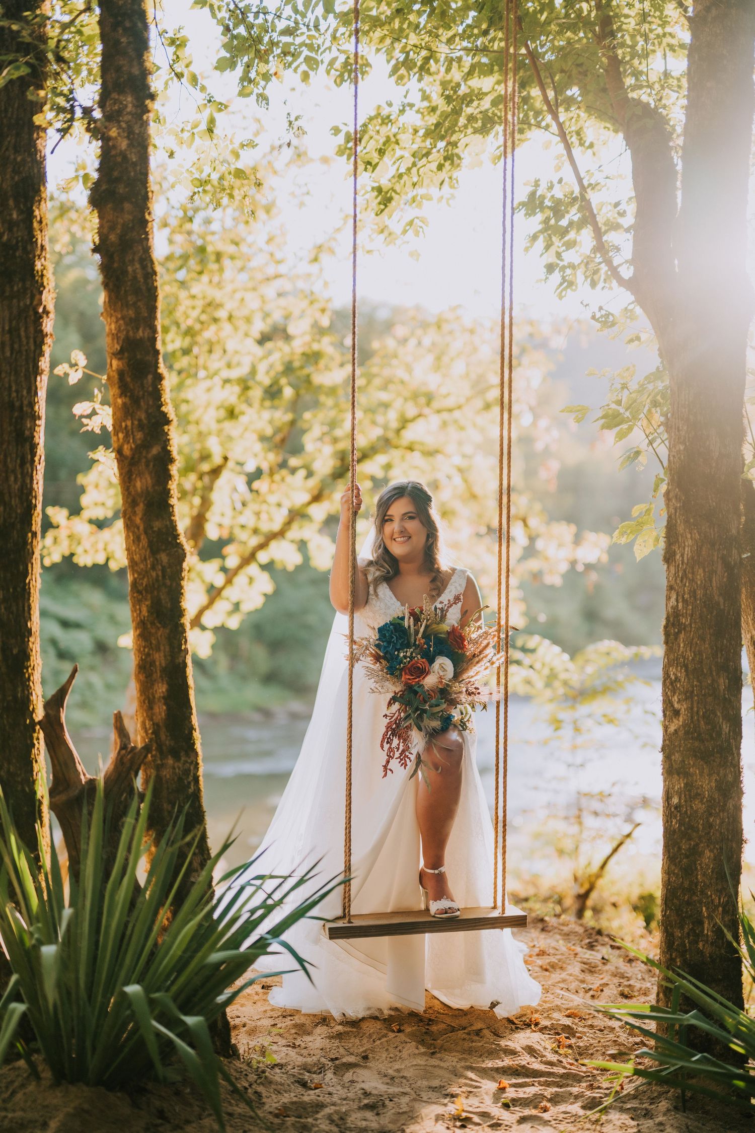 Bride in white dress poses on wooden swing in sunlit garden with trees and greenery.