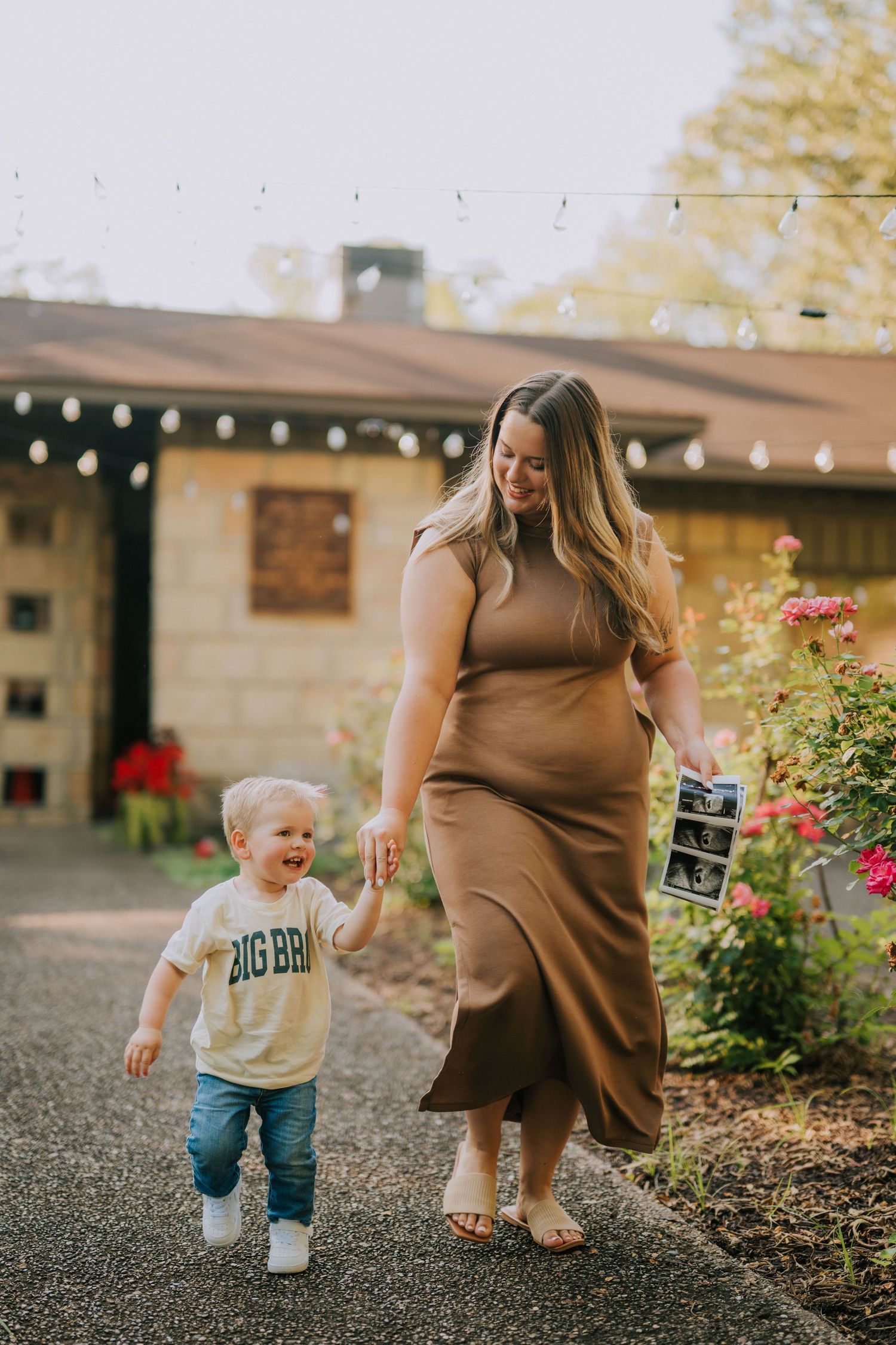 A toddler in casual clothes runs near garden flowers while someone holds a sonogram photo.