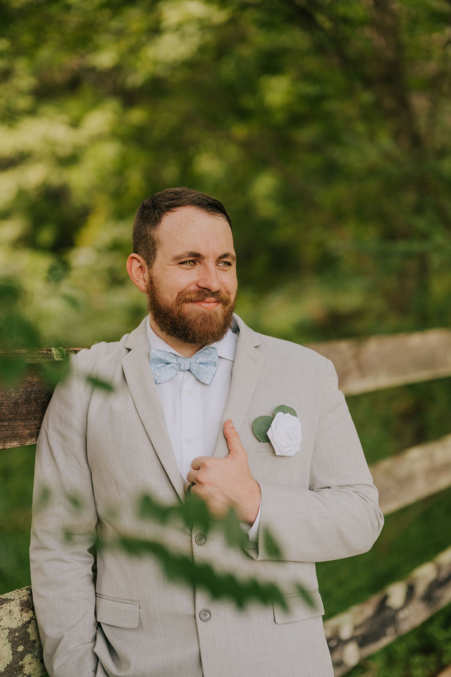 A groom in a light gray suit and white boutonniere smiles while adjusting his tie outdoors.